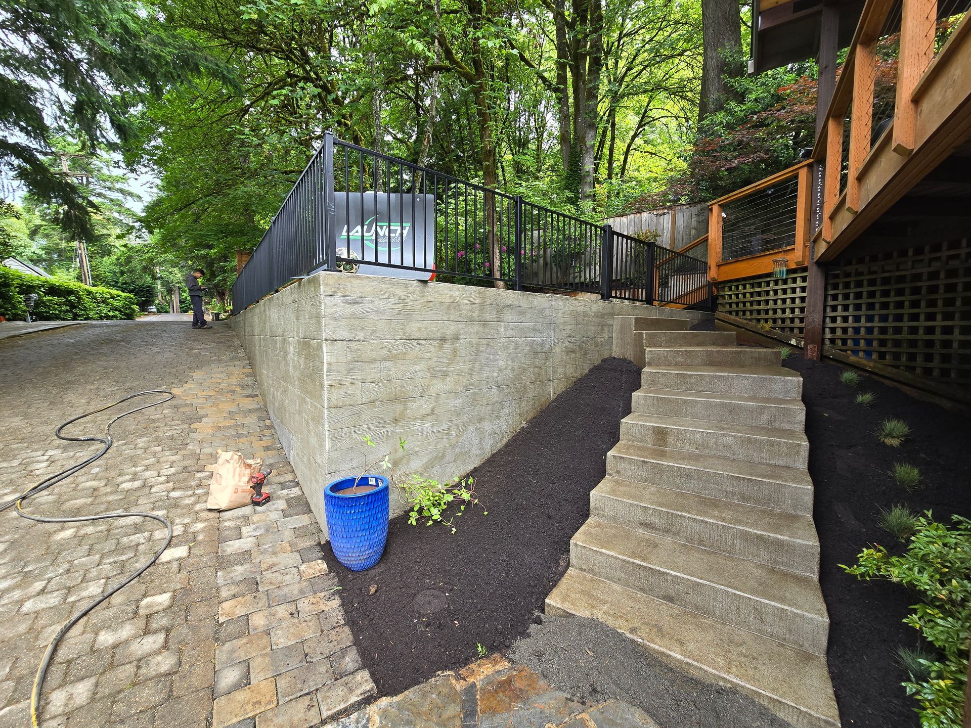 Steep outdoor steps beside a stone retaining wall and a blue bucket, surrounded by trees and a wooden deck