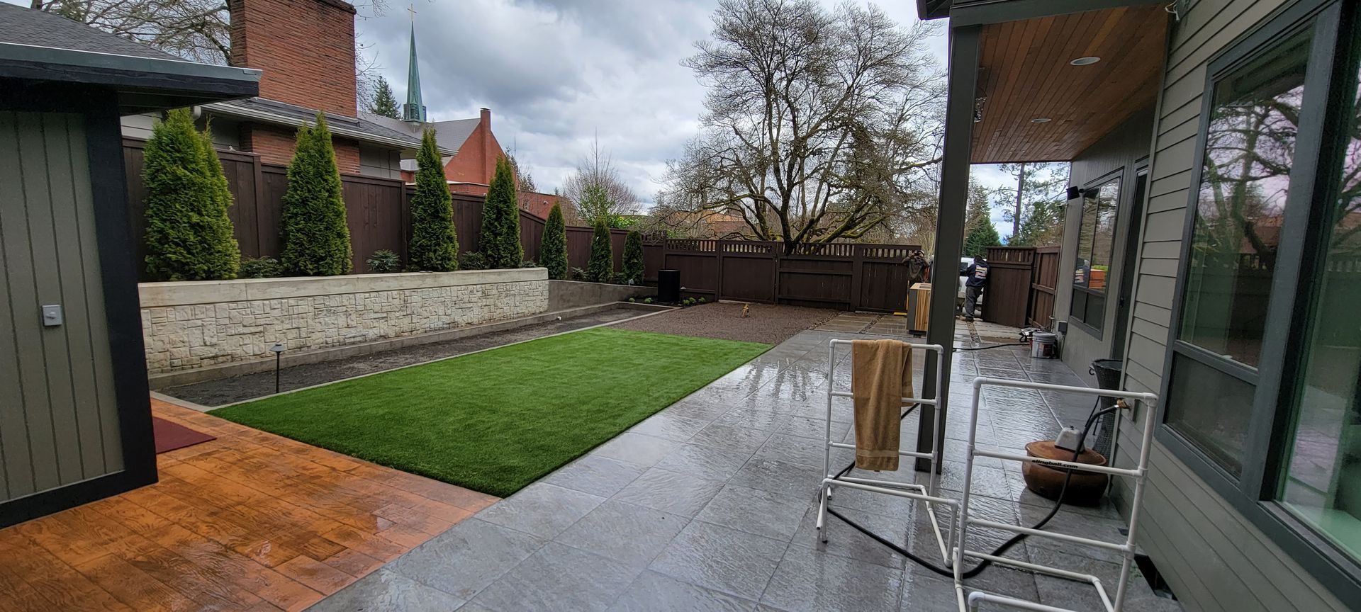 Backyard patio with wet stone paving, green lawn, brick wall, and outdoor seating under a cloudy sky