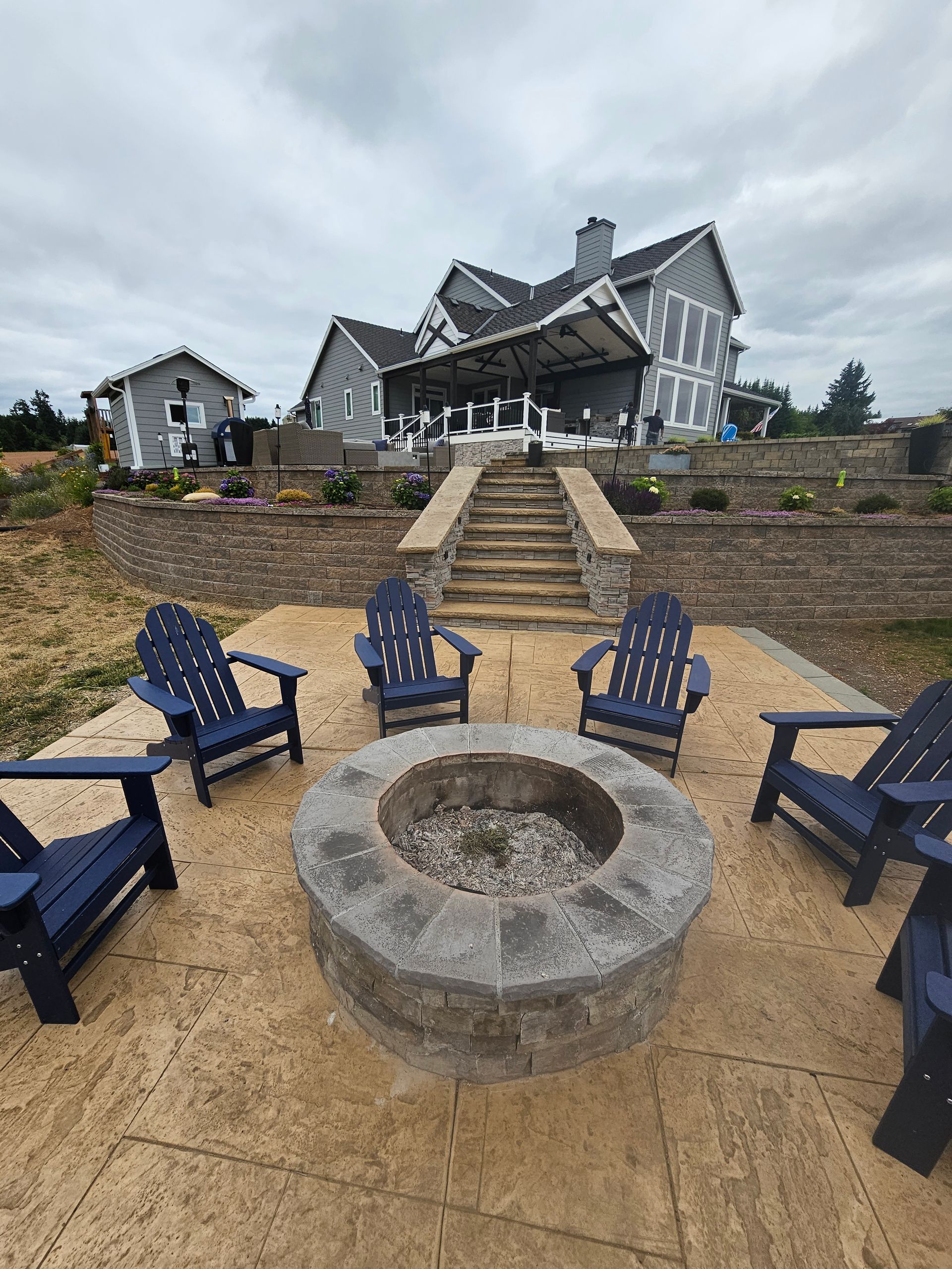 Stone fire pit surrounded by blue chairs on a sandy patio before a large gray house under cloudy skies