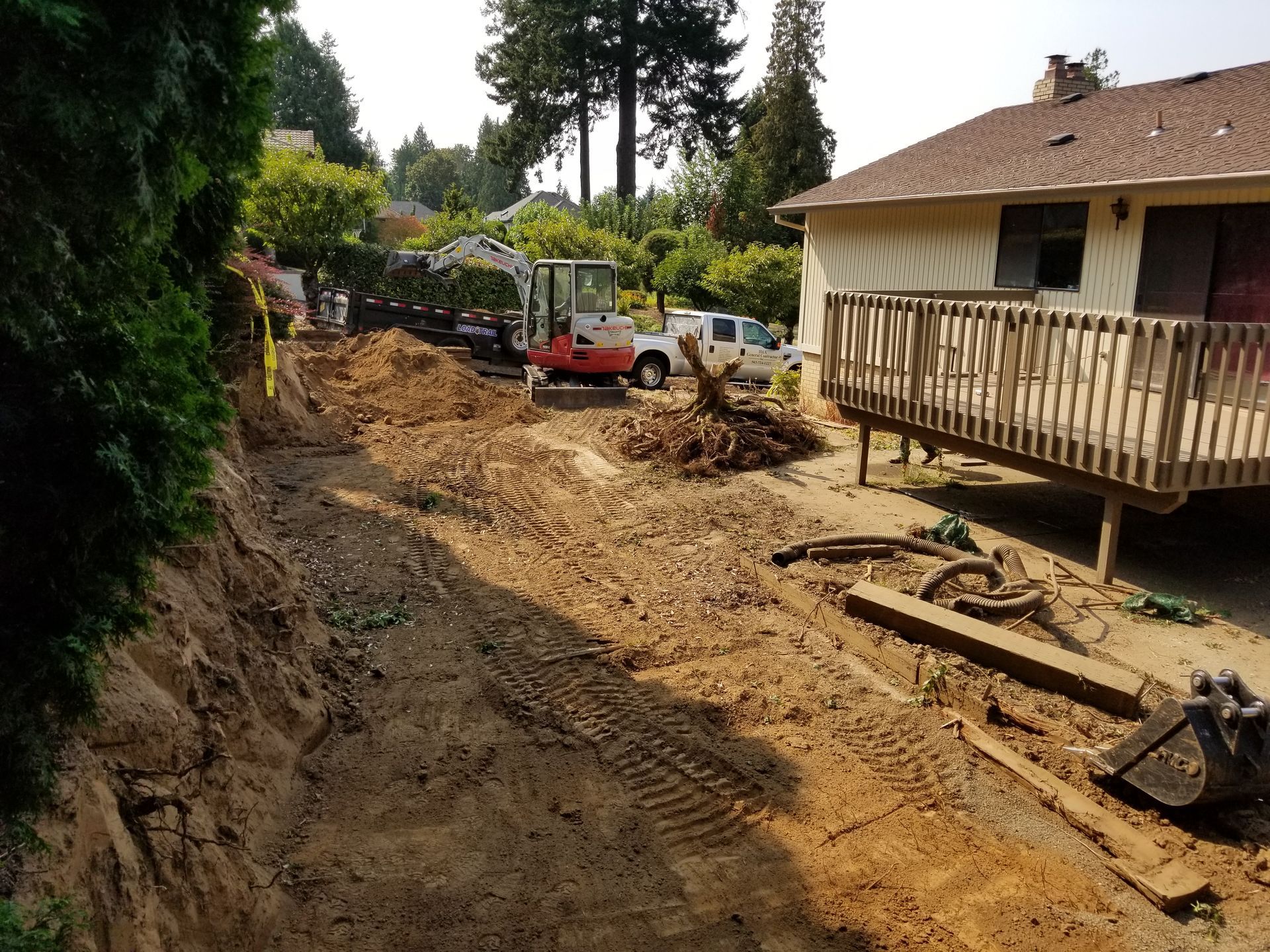 Mud-filled backyard construction beside a house and deck, with an excavator and dirt piles.
