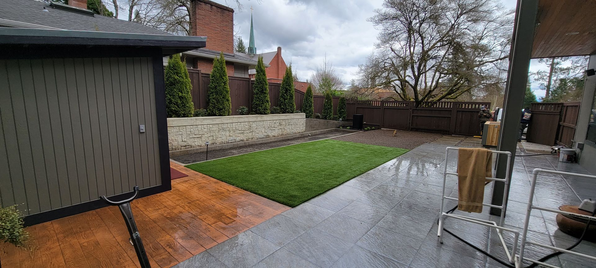 Backyard patio with wet stone, wooden deck, green lawn, shed, and leafless trees on a rainy day
