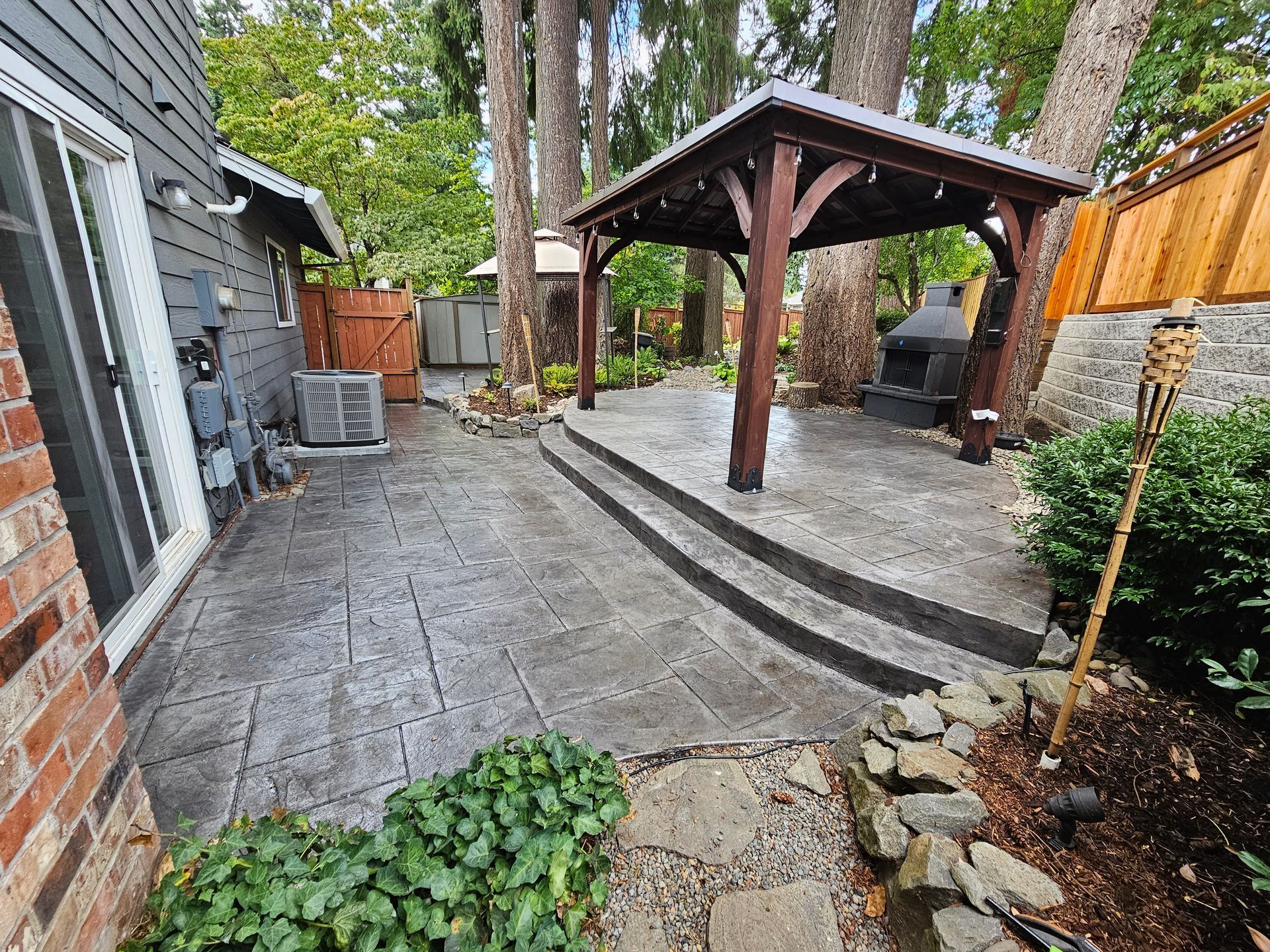 Backyard patio with a wooden gazebo, curved stone steps, and trees along a fenced yard.