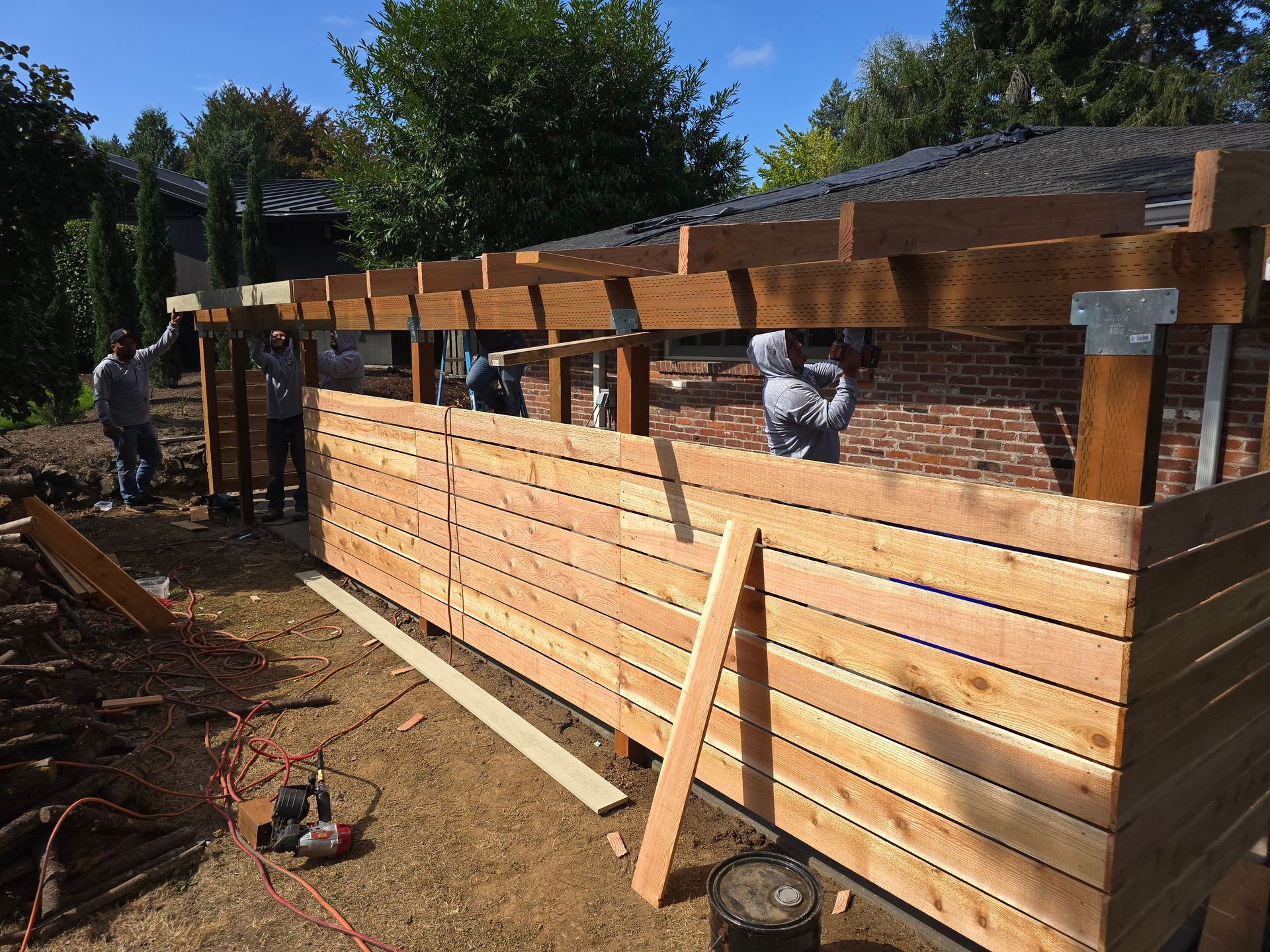 People building a wooden fence beside a brick house in a sunny yard