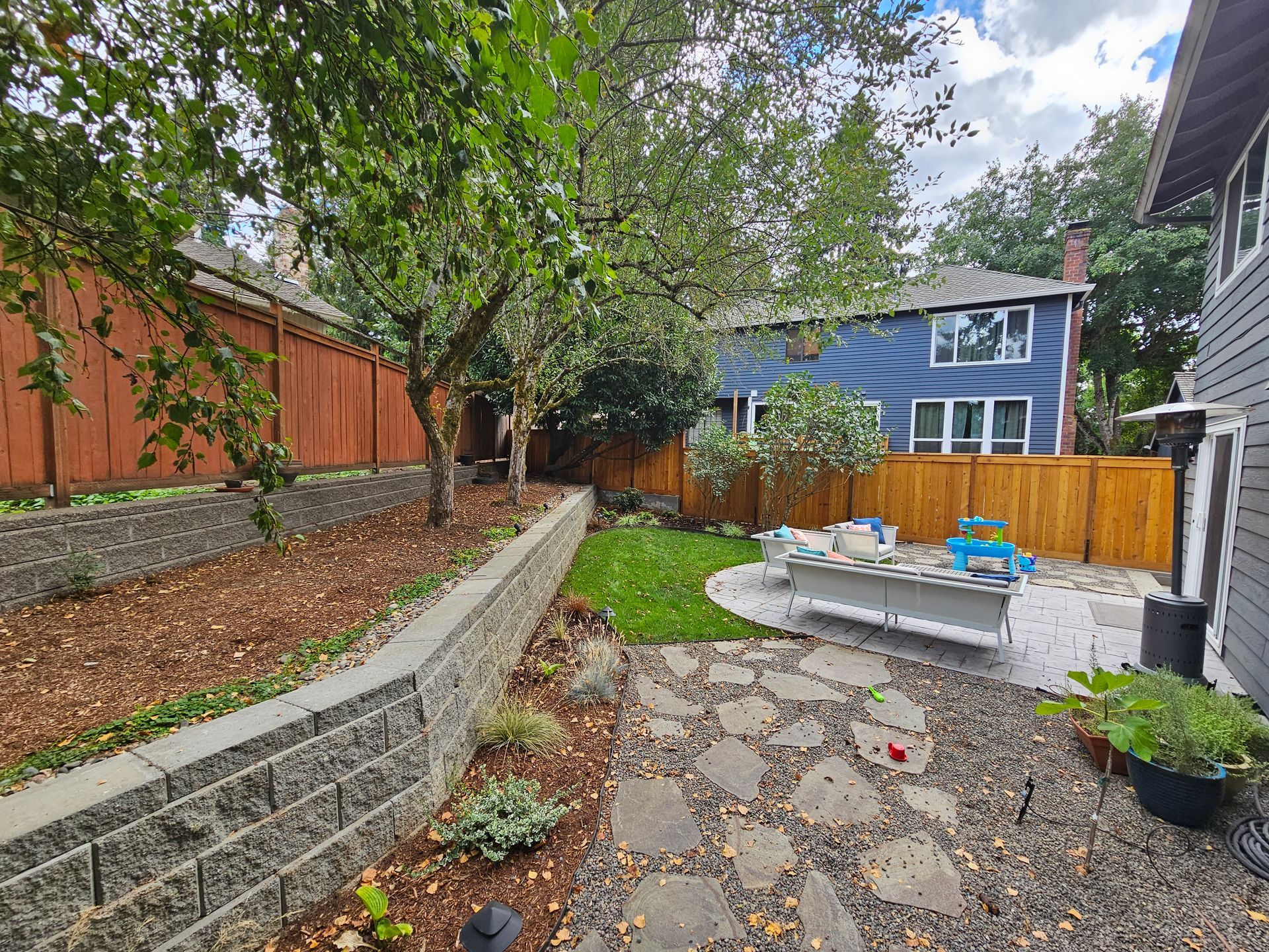Backyard patio with stone path, raised garden bed, trees, and a blue house beyond the fence