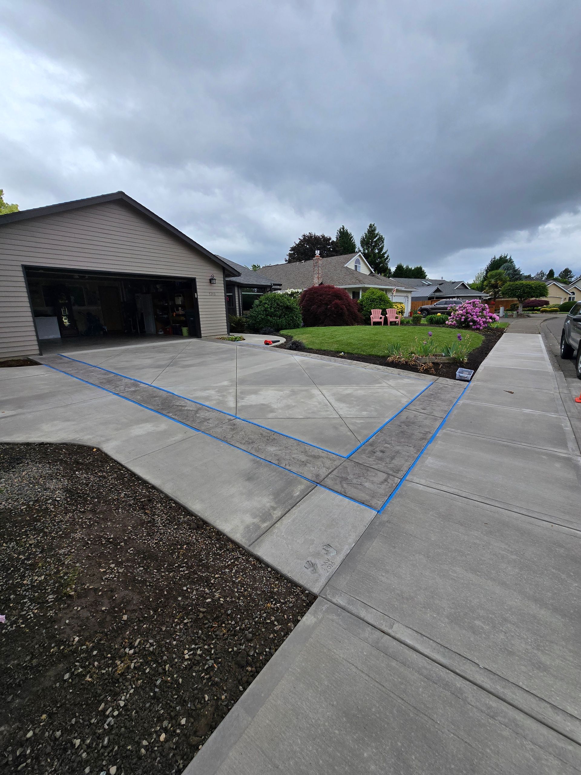 Suburban driveway and garage under cloudy sky, with flower beds and concrete walkway.