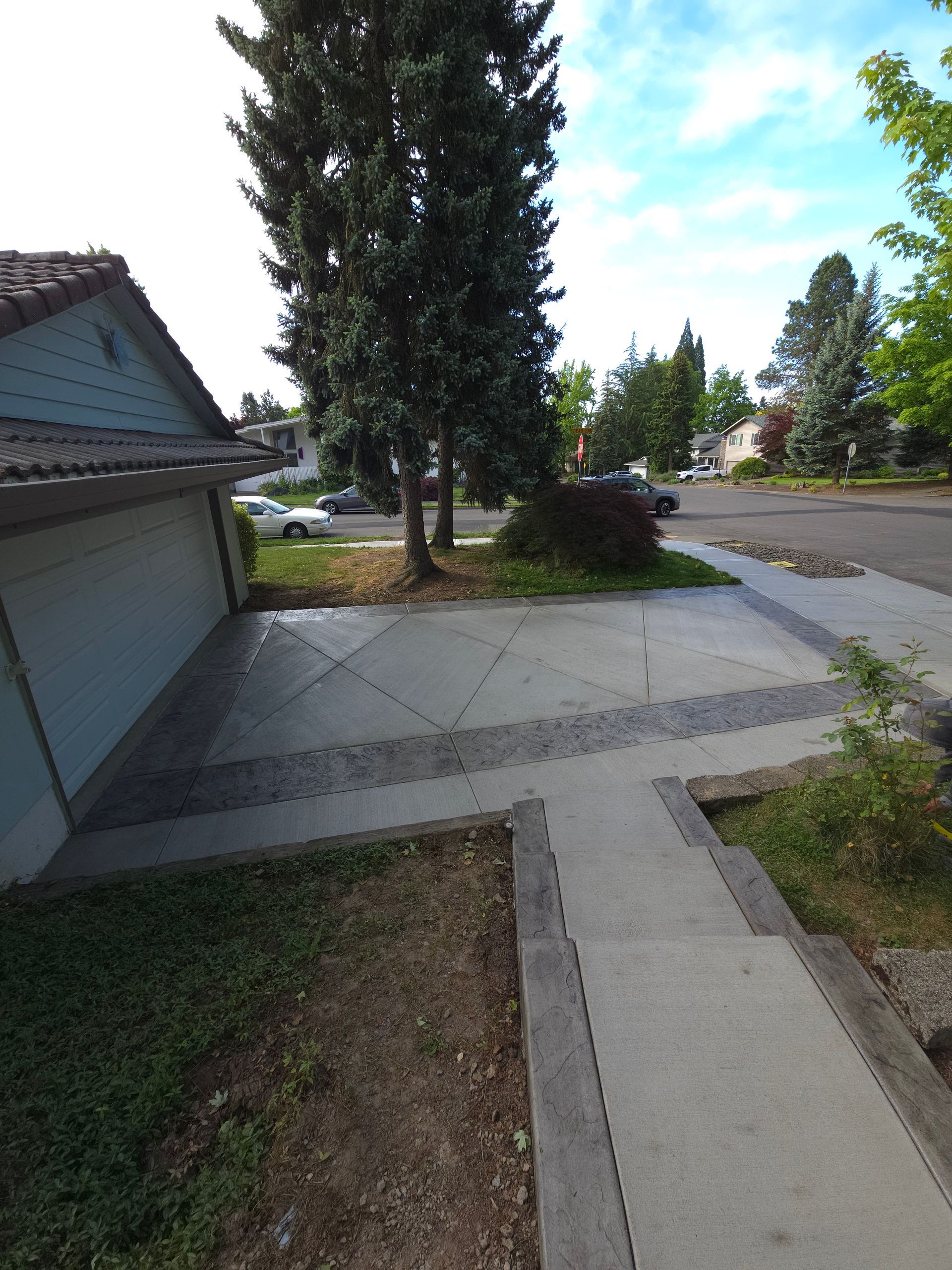 Suburban driveway and walkway beside a garage, with tall trees, shrubs, and a quiet street.