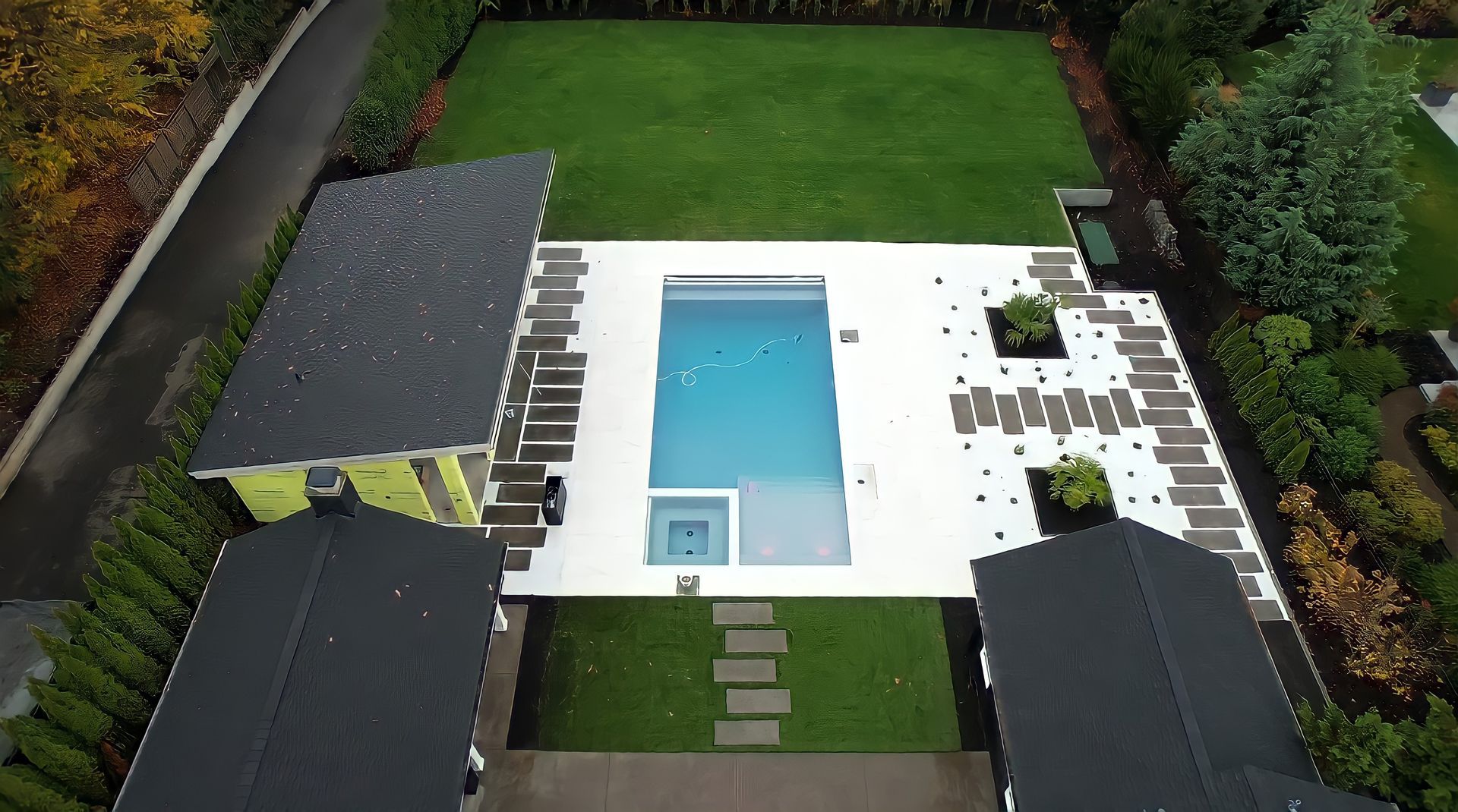 Aerial view of a backyard with a rectangular pool, white deck, lounge chairs, and green lawn.