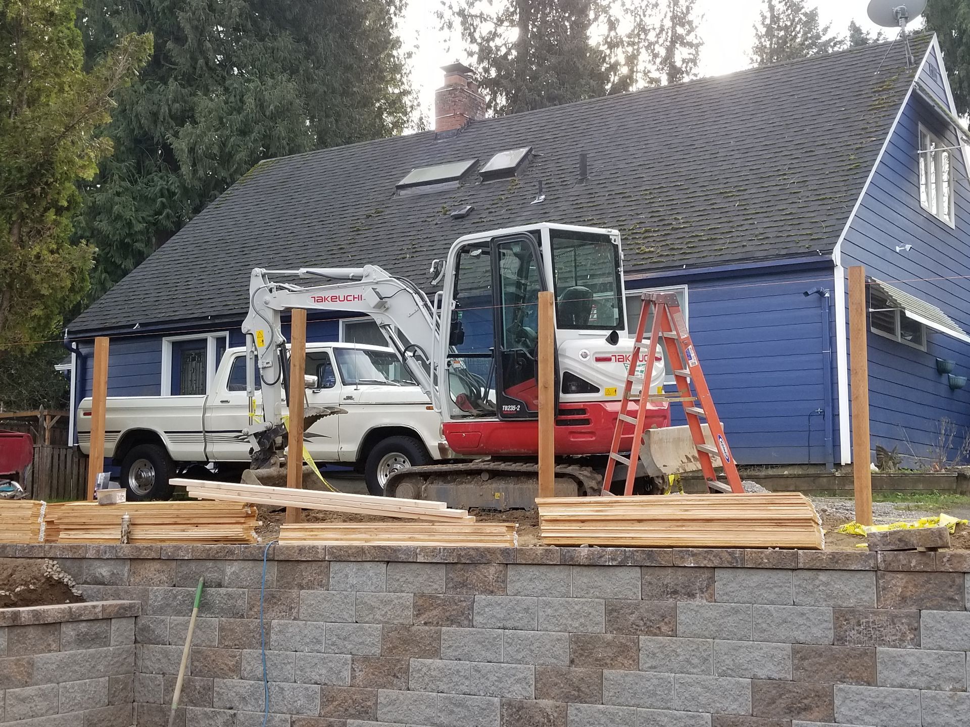House renovation with excavator, white truck, and lumber in front of a blue building