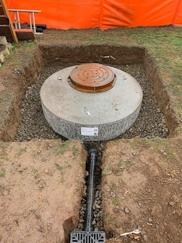 Circular concrete utility chamber in a dug trench, with a rust-colored cover and orange safety barrier behind it