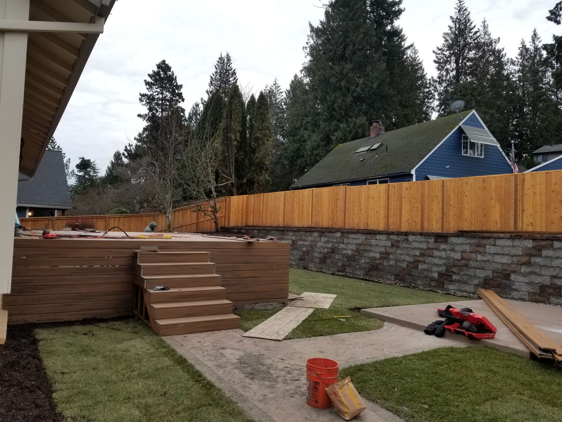 Backyard with wooden deck stairs, stone retaining wall, lawn, and a red toy car near a fence.