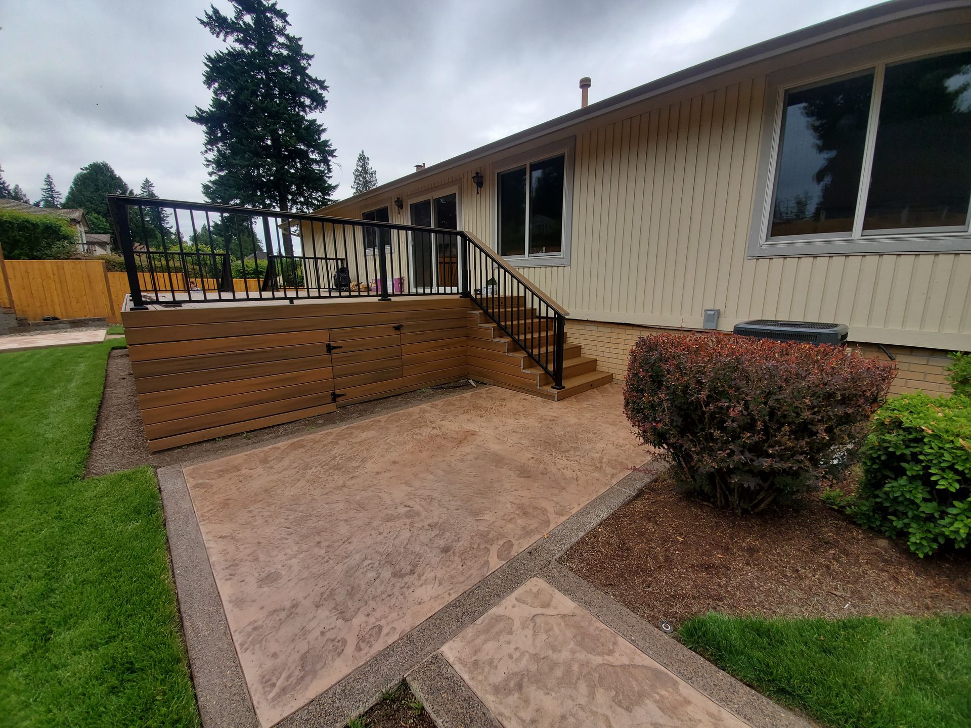 Backyard patio with a wooden deck, steps, black railing, and brick retaining wall beside a house