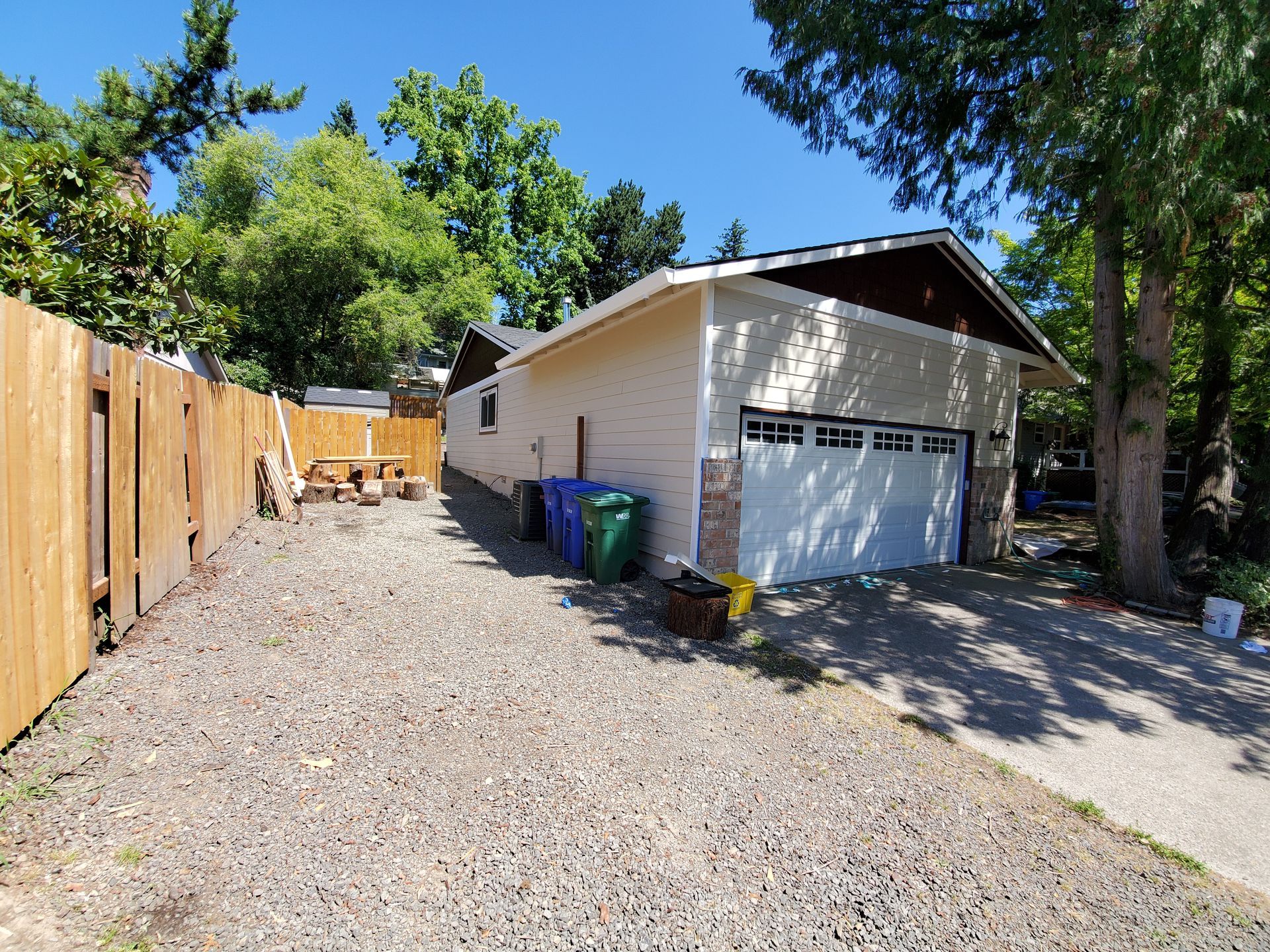Driveway beside a wooden fence leading to a white garage with gravel and trees on a sunny day
