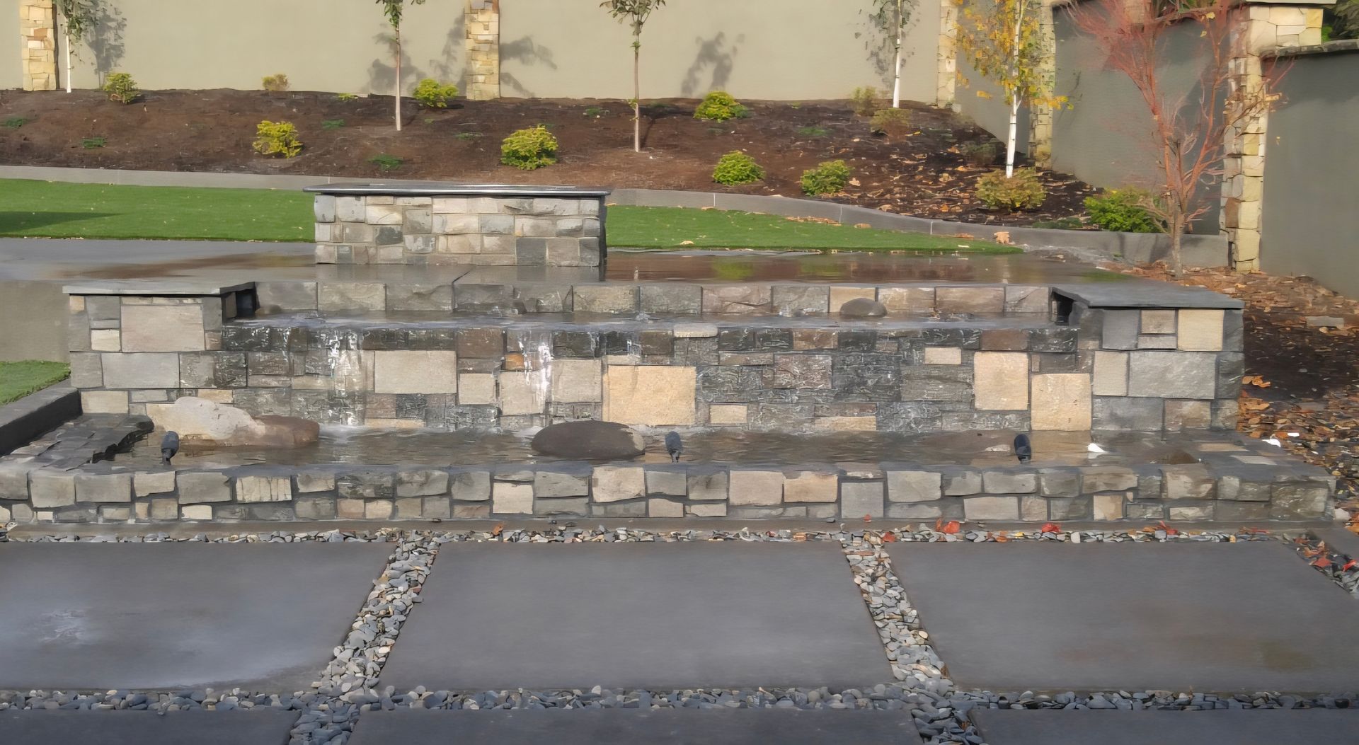 Stone retaining wall with steps in front of a landscaped garden bed beside a paved driveway.