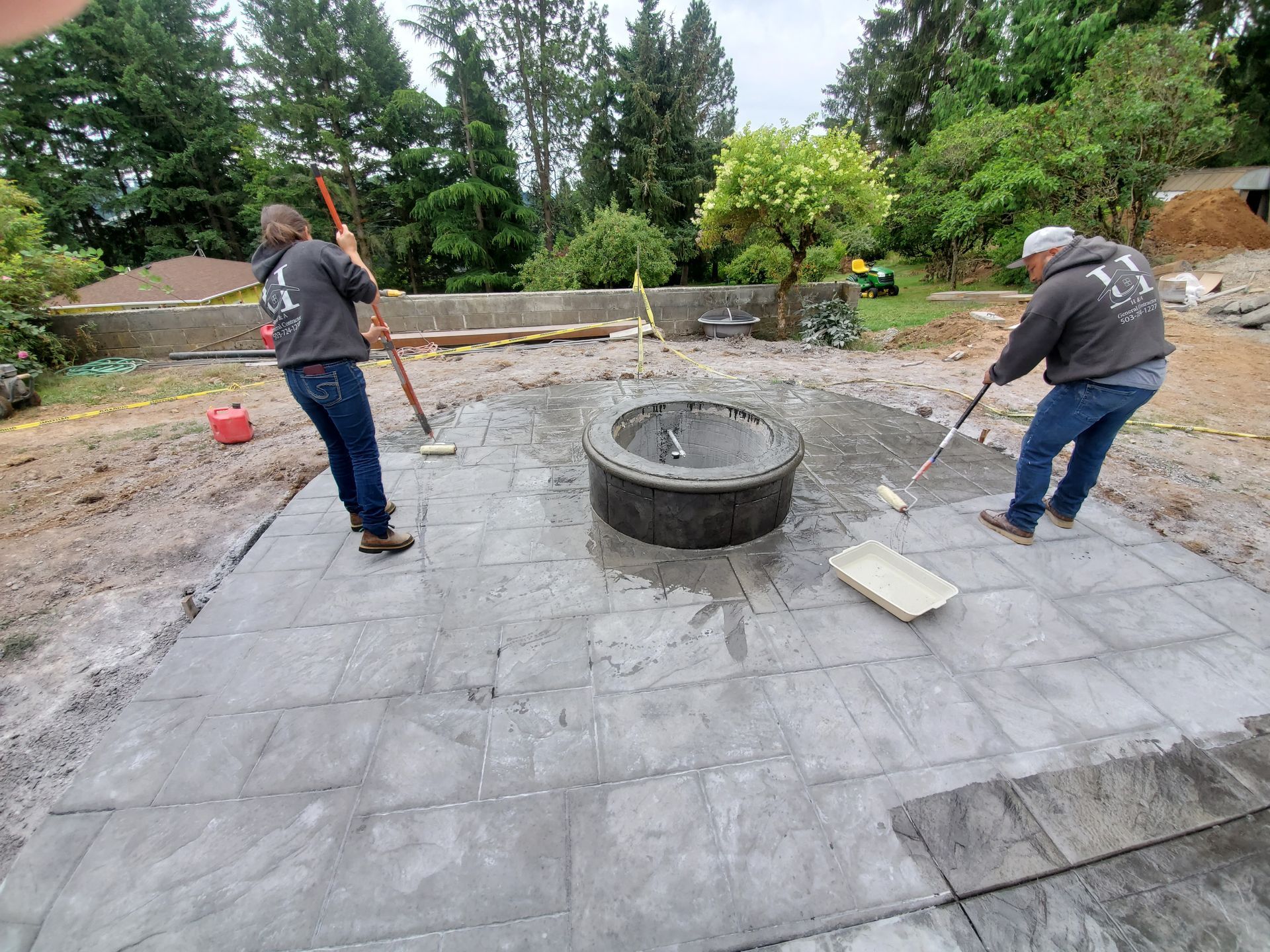 Two workers repairing a stone patio around a circular fire pit in a backyard garden
