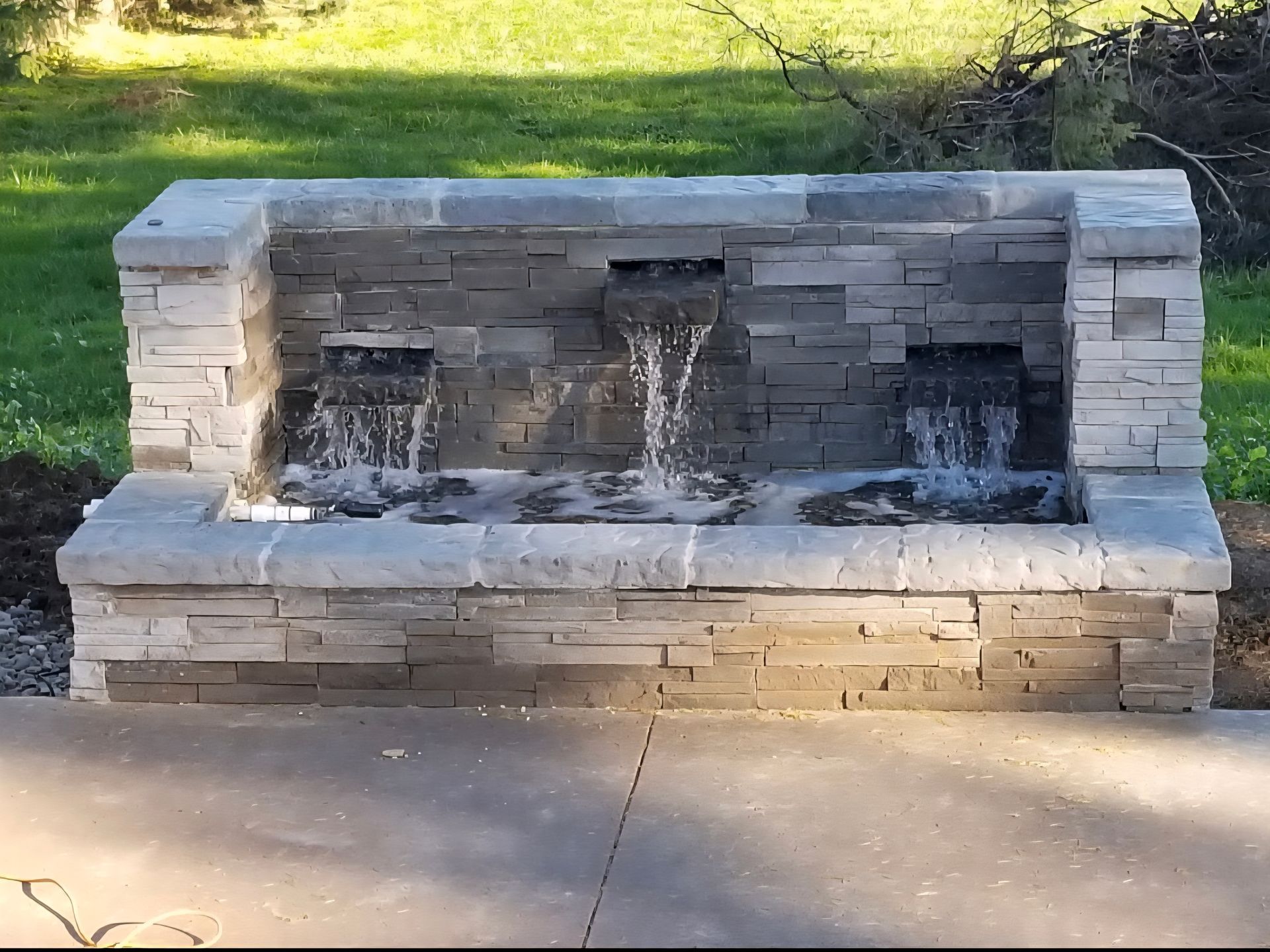 Stone outdoor fountain with multiple water streams in a park, set against grass and a paved walkway