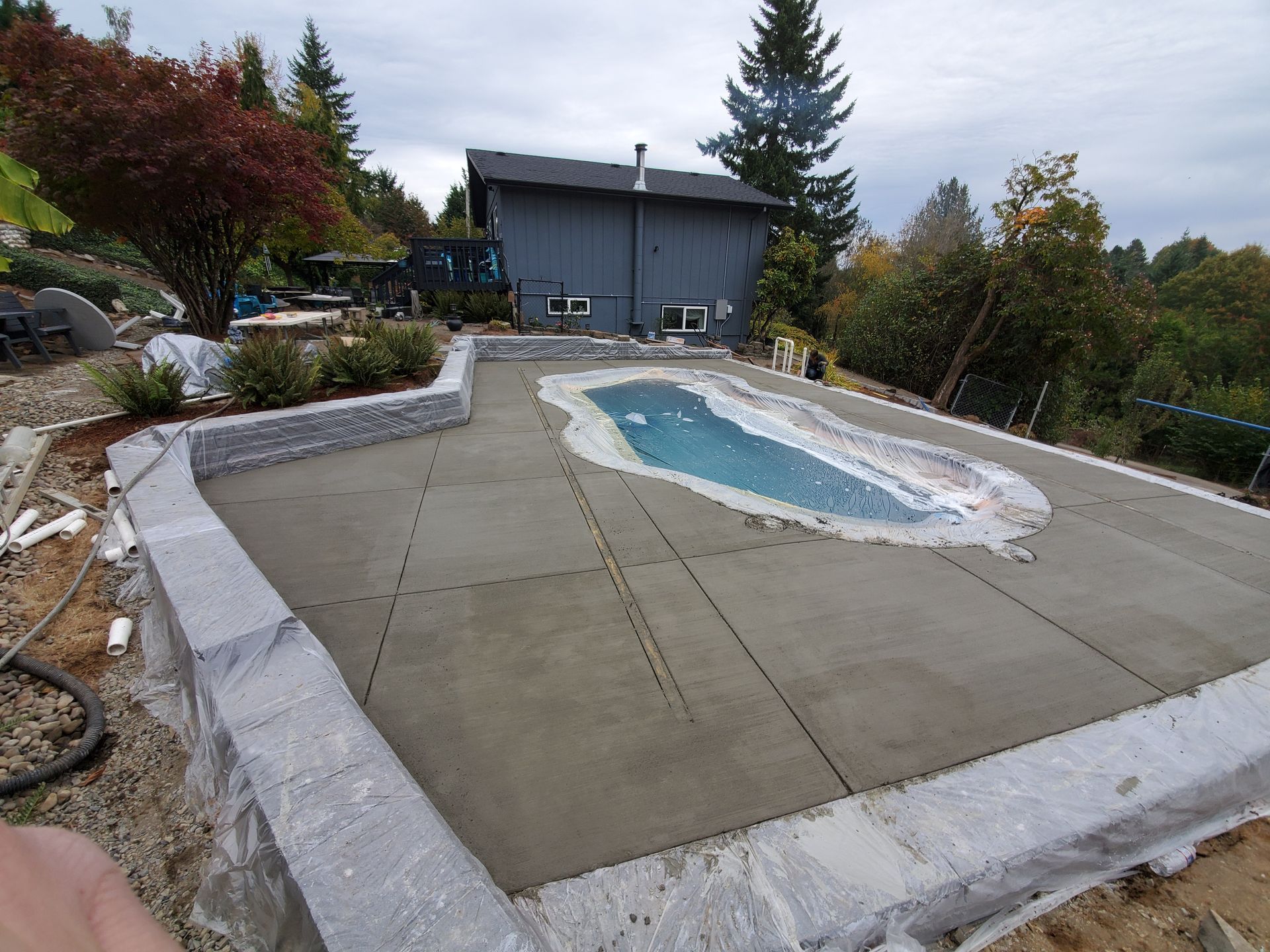 Empty backyard concrete pool deck with an unfinished in-ground pool beside a gray house.