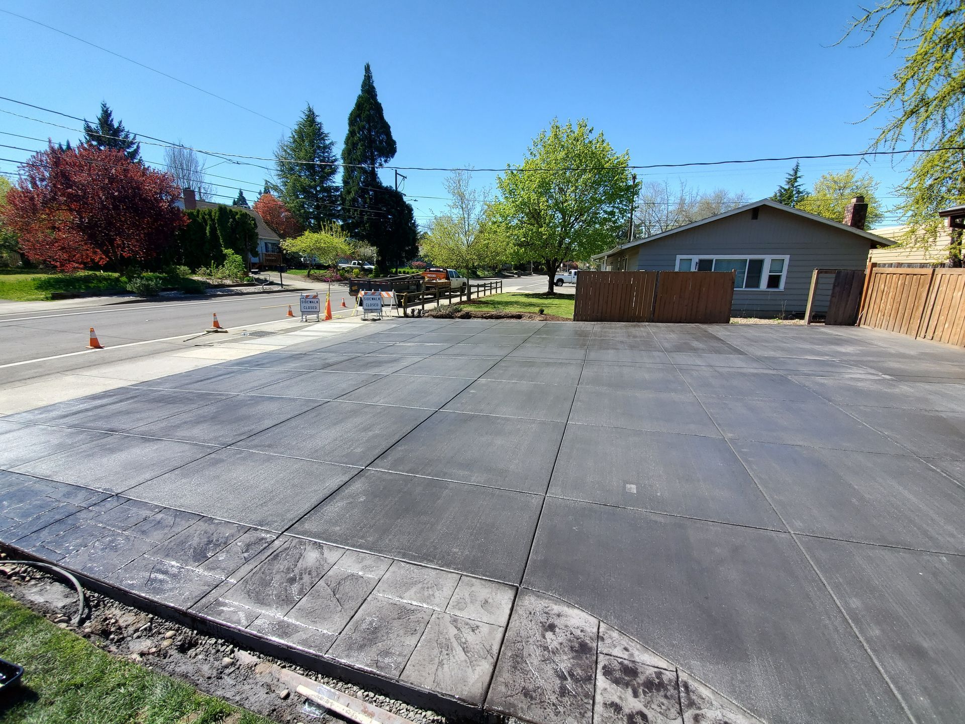 Empty paved lot beside a house and trees under a clear blue sky.