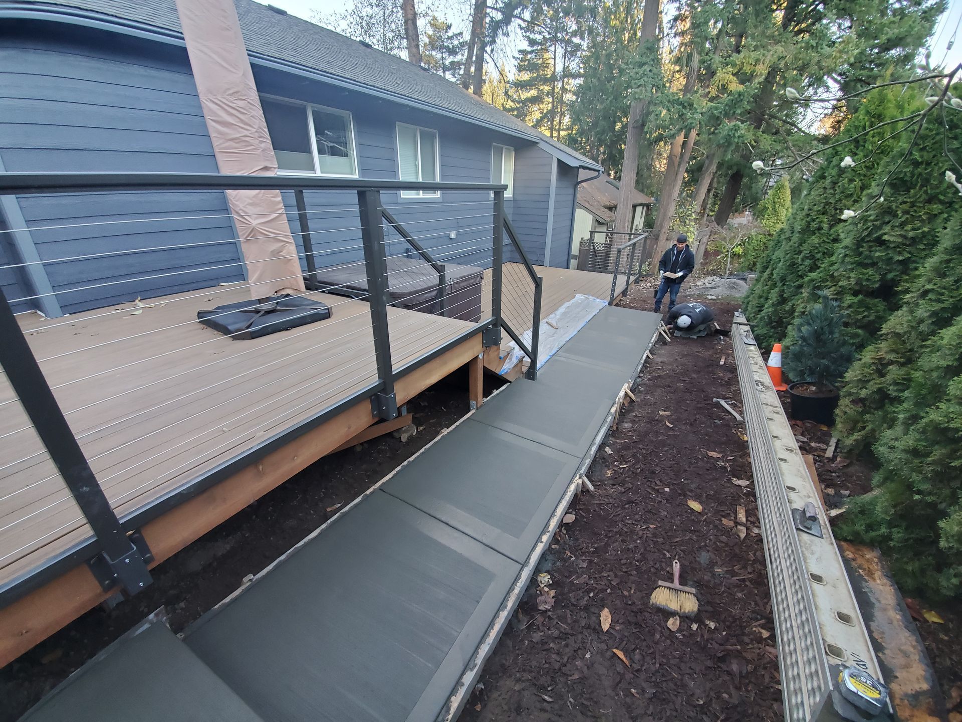 Backyard deck ramp with black railings leading to a walkway beside a house and garden bed