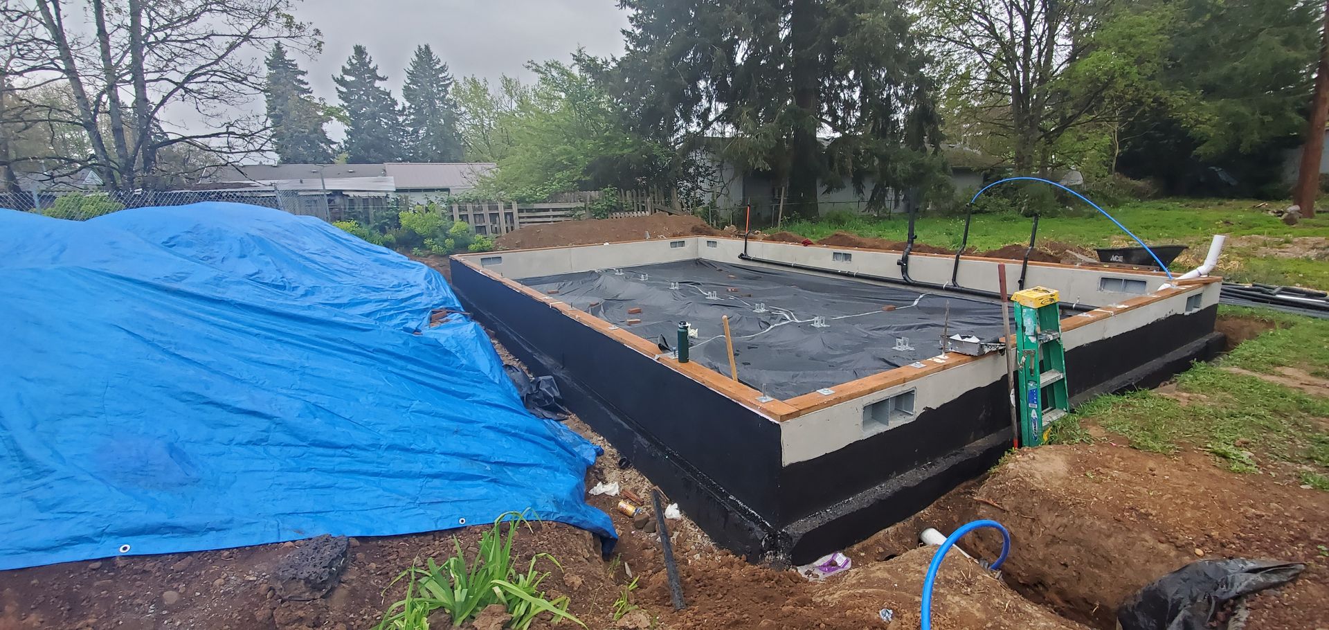 Construction site with a concrete foundation, blue tarp, and worker near the corner in a wooded yard