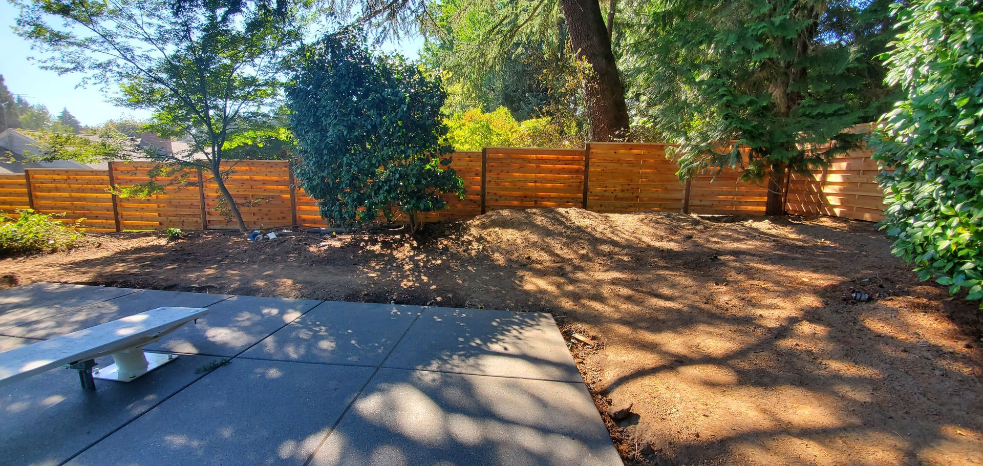 Backyard patio with a wooden fence, trees, and dappled sunlight over mulch and paving stones