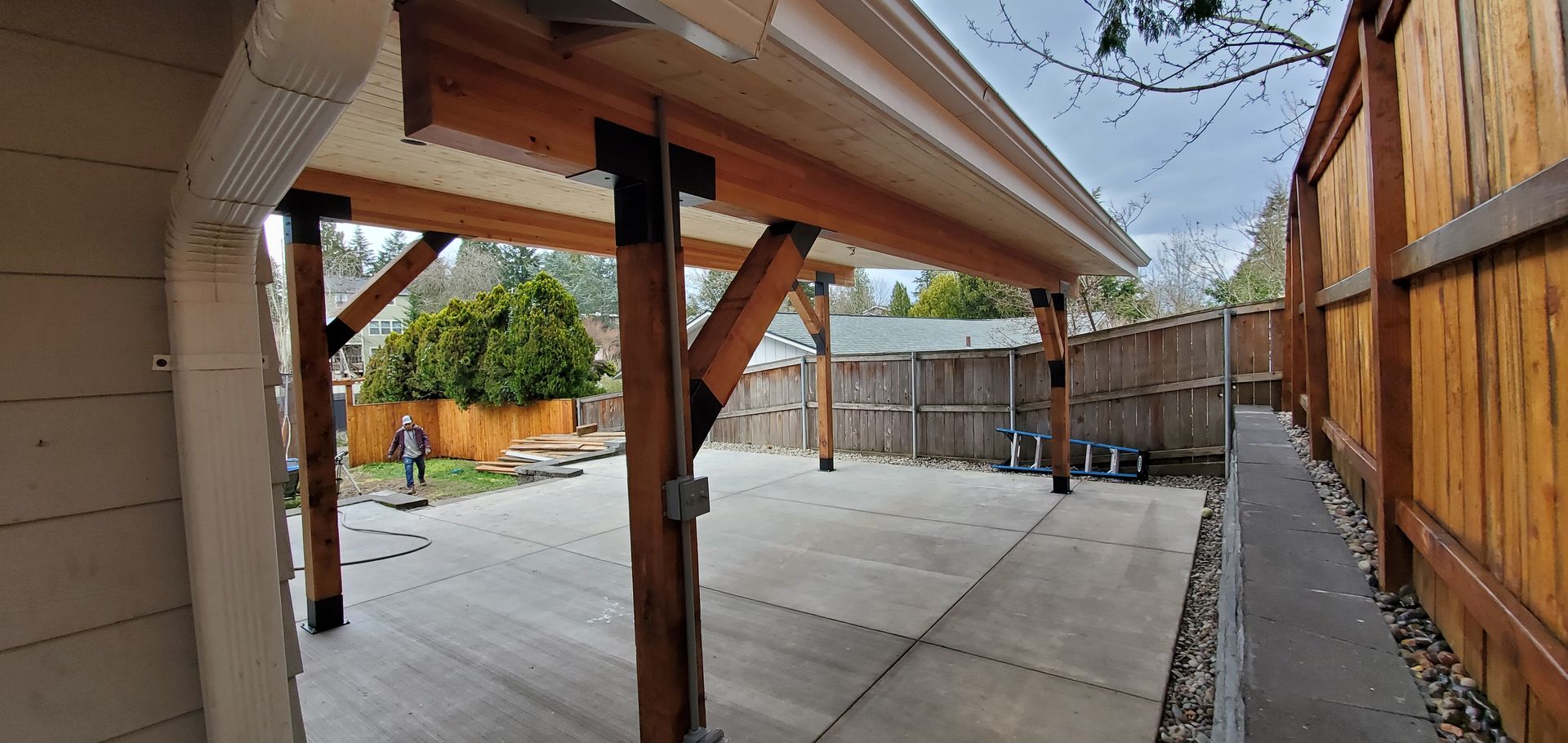 Covered concrete patio with wooden posts, fence, and a small person in the distance under a bright sky