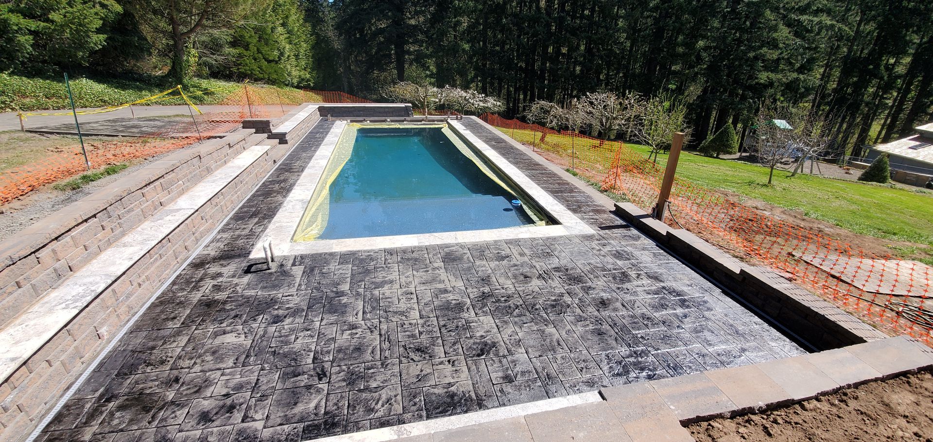 Rectangular outdoor swimming pool under construction, bordered by gravel and brickwork in a wooded area