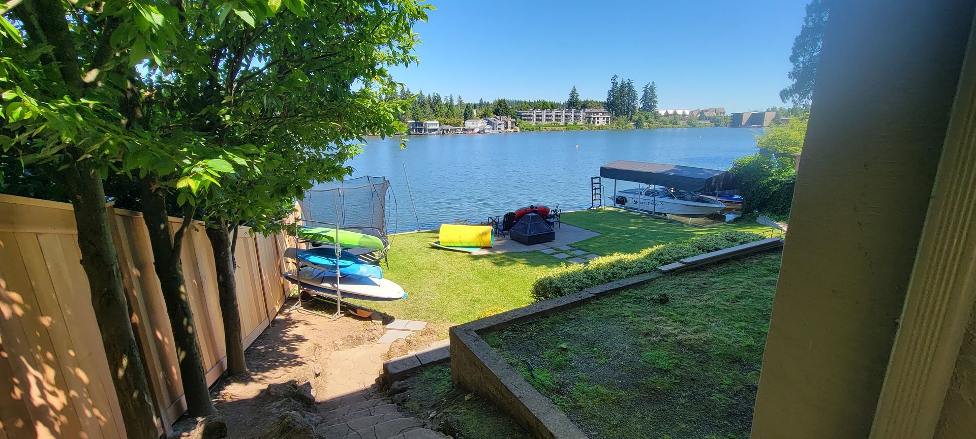Sunny lakeside yard with wooden fence, grassy slope, small boat, and dock across the water