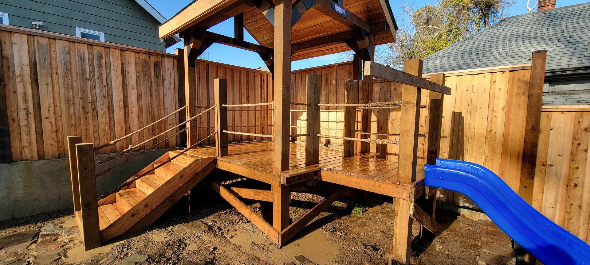 Wooden backyard playset with platform, roof, benches, and blue slide in a fenced yard