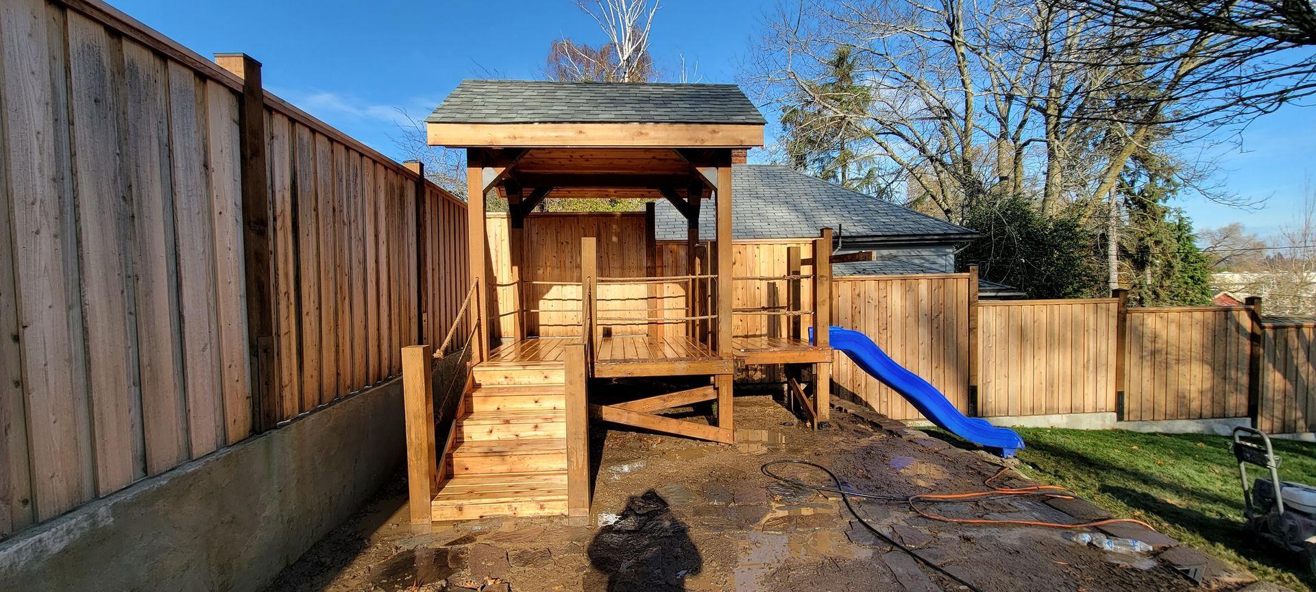 Wooden backyard playhouse with stairs and a blue slide in a fenced yard