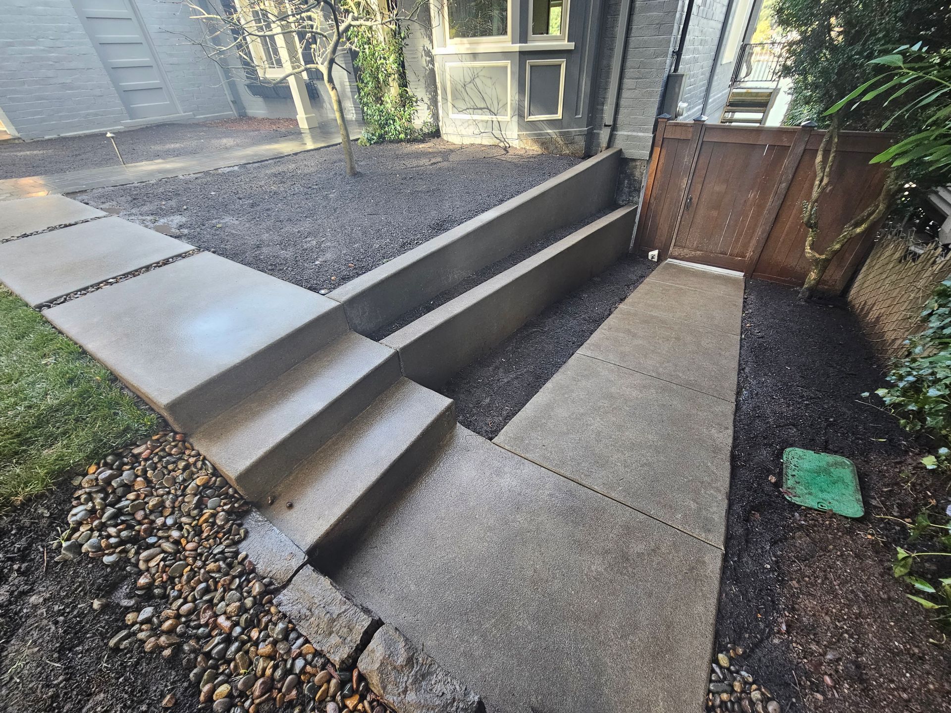 Concrete outdoor steps and ramp beside a house, bordered by gravel and a wooden gate