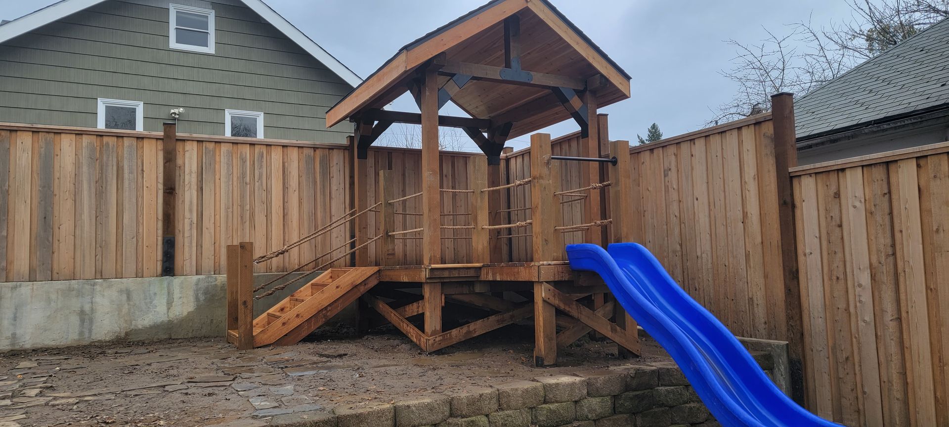 Wooden backyard playset with a blue slide and roofed platform beside a fenced yard.