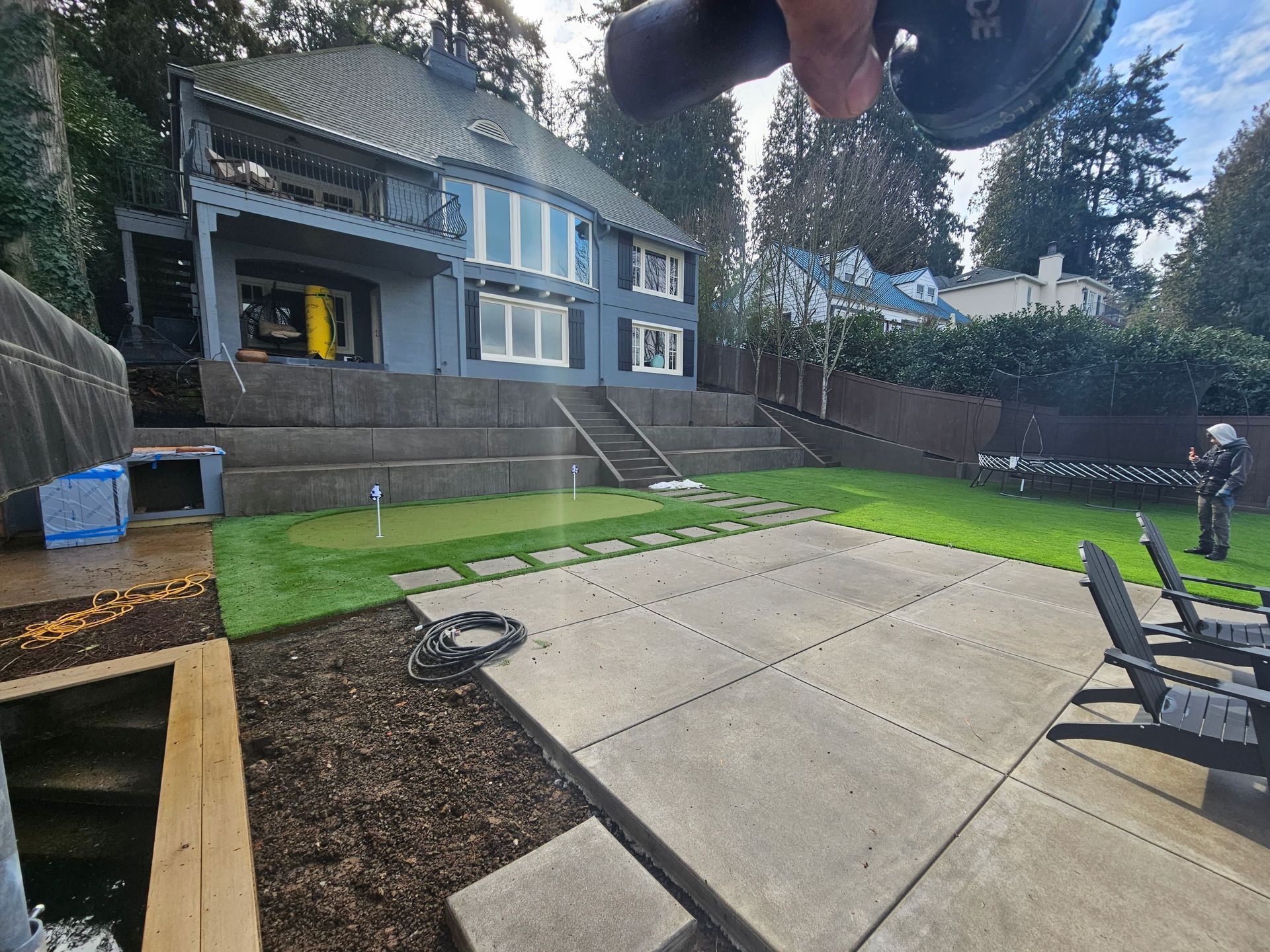 Backyard patio with concrete slabs, lawn, wooden deck, and a house with stairs and windows.