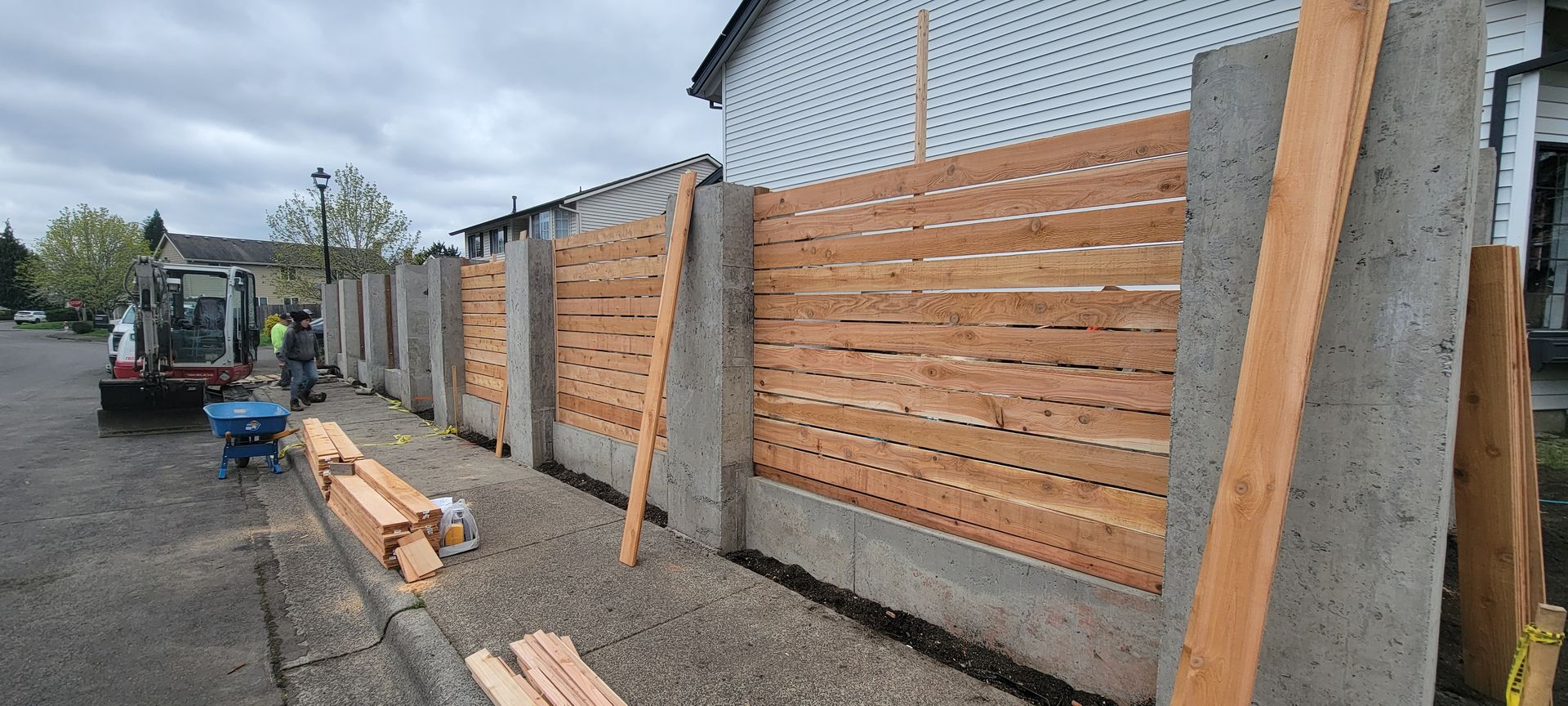 Wooden fence panels being installed along a sidewalk beside a road, with stacked lumber and a worker on a loader.