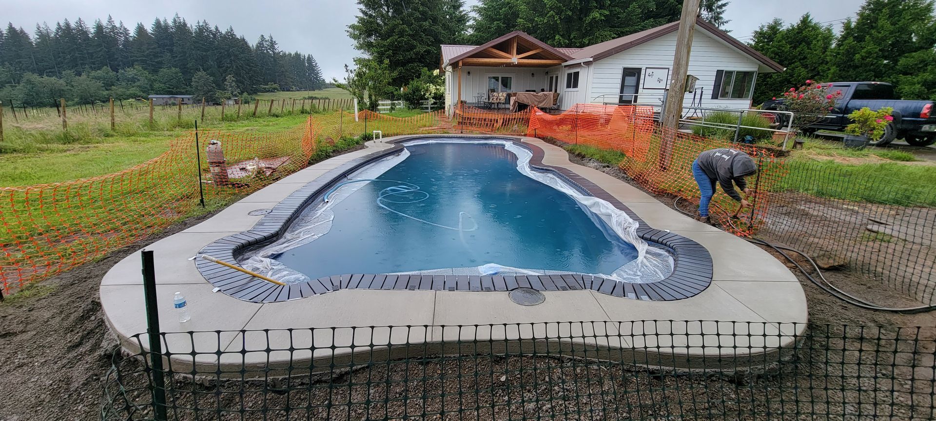 Above-ground pool under construction beside a house, with workers and orange safety fencing in a yard