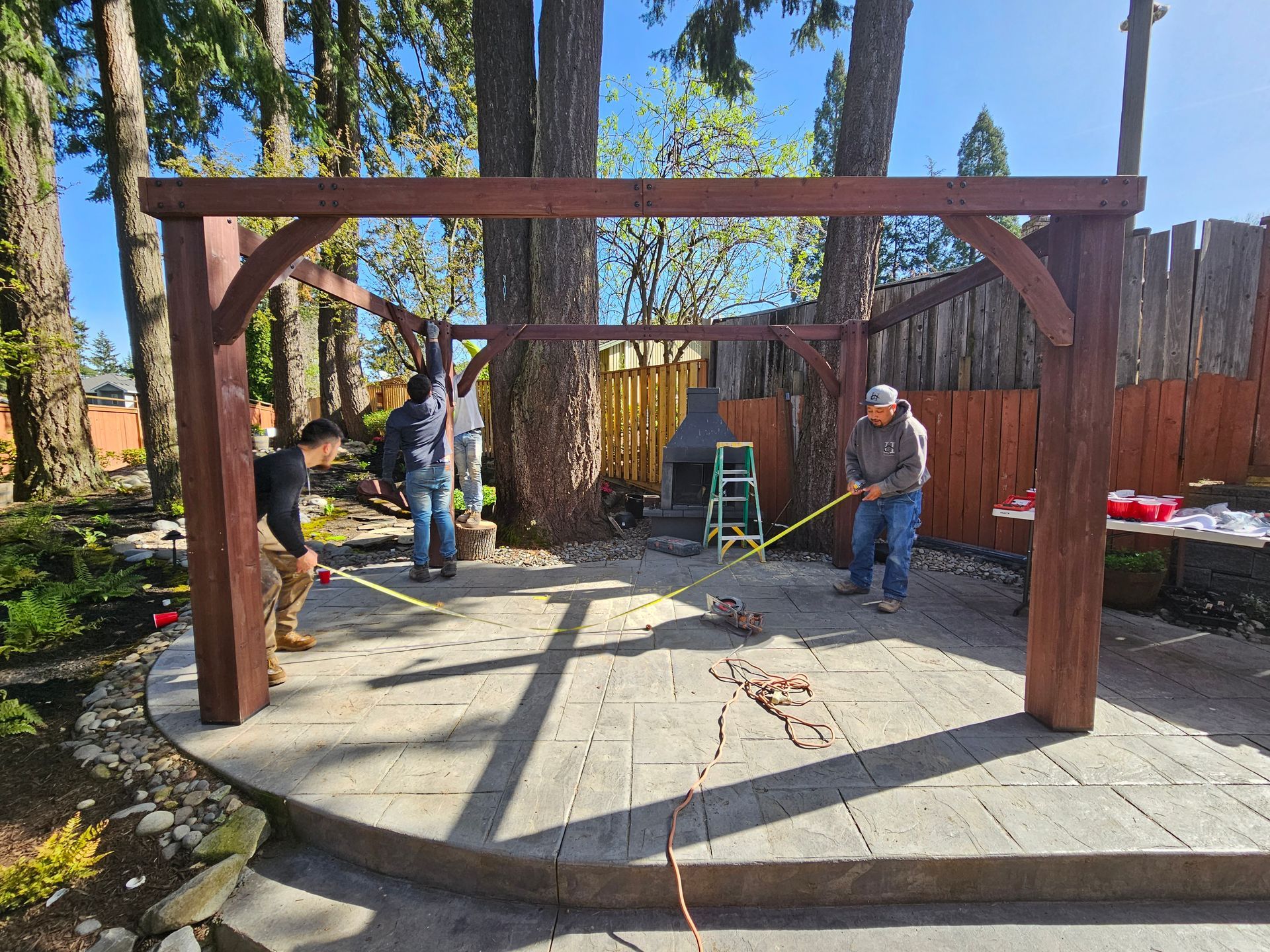 People installing a large wooden arch frame on a patio in a backyard.