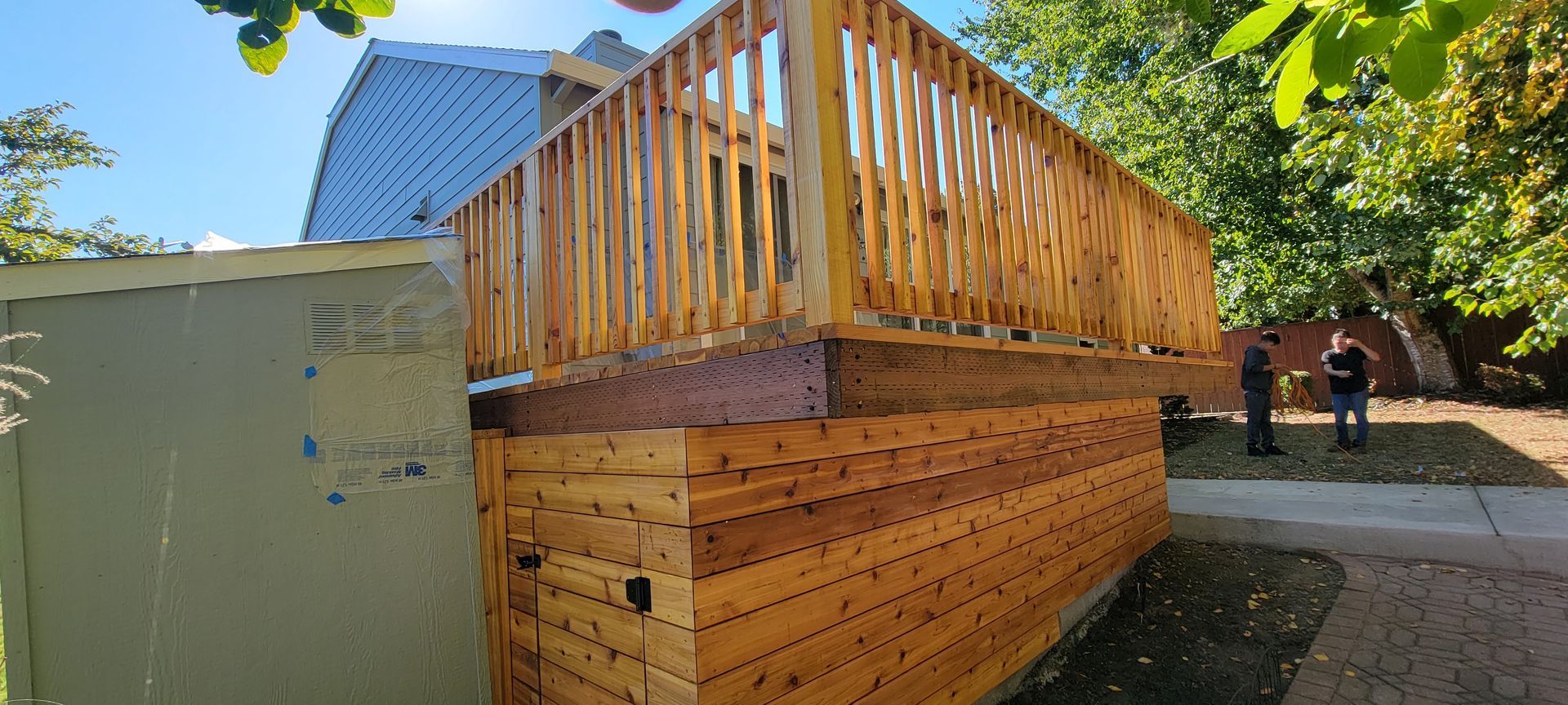 Wooden deck and raised platform beside a green building, with two people in the background.