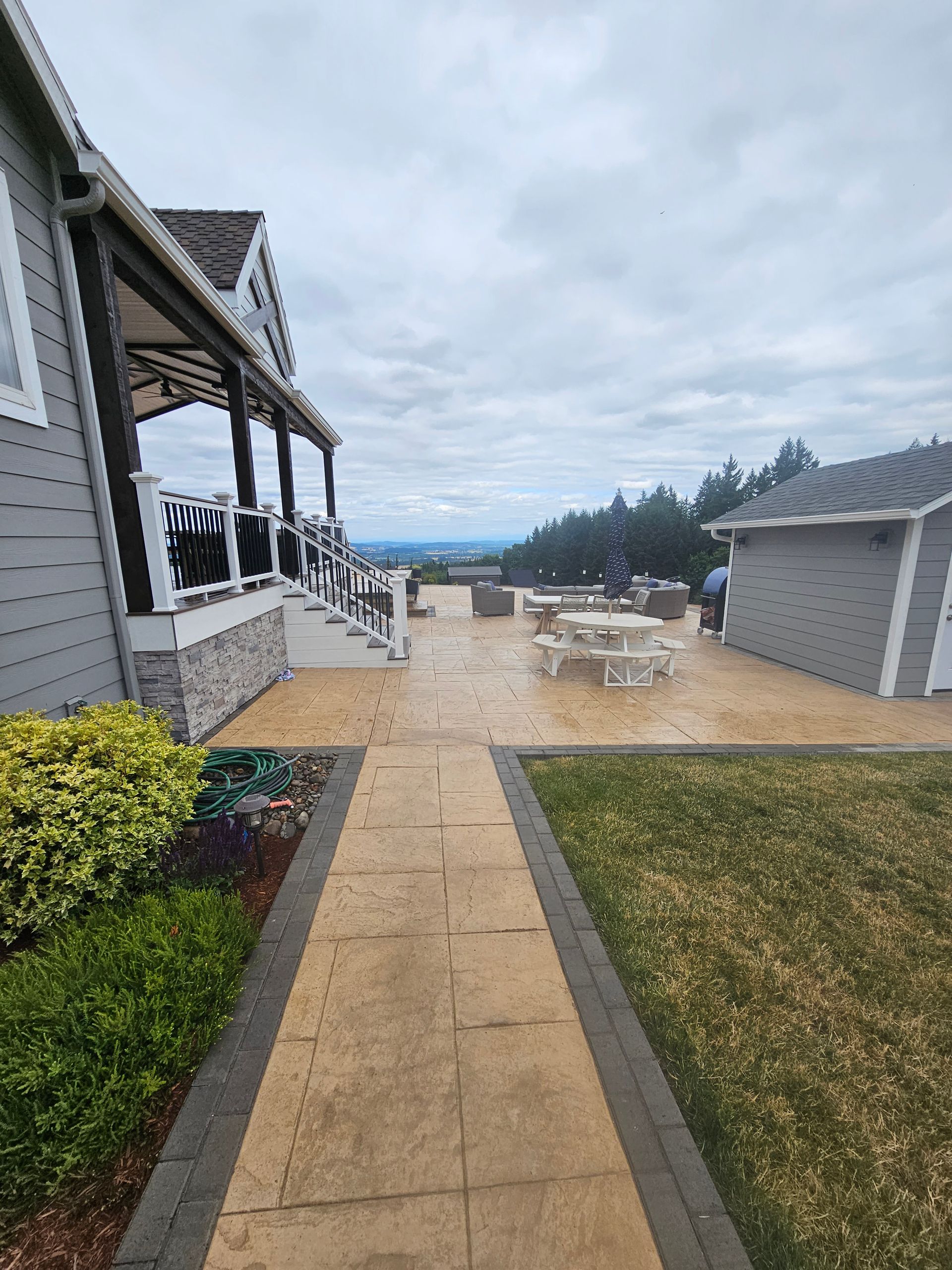 Wet stone walkway beside gray houses leading to a patio and distant ocean under cloudy skies
