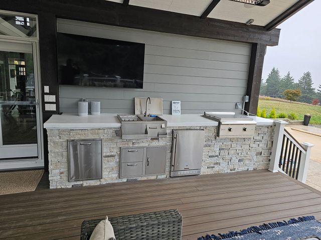 Outdoor patio kitchen with built-in grill, stone counter, sink, and wall-mounted TV under a covered deck