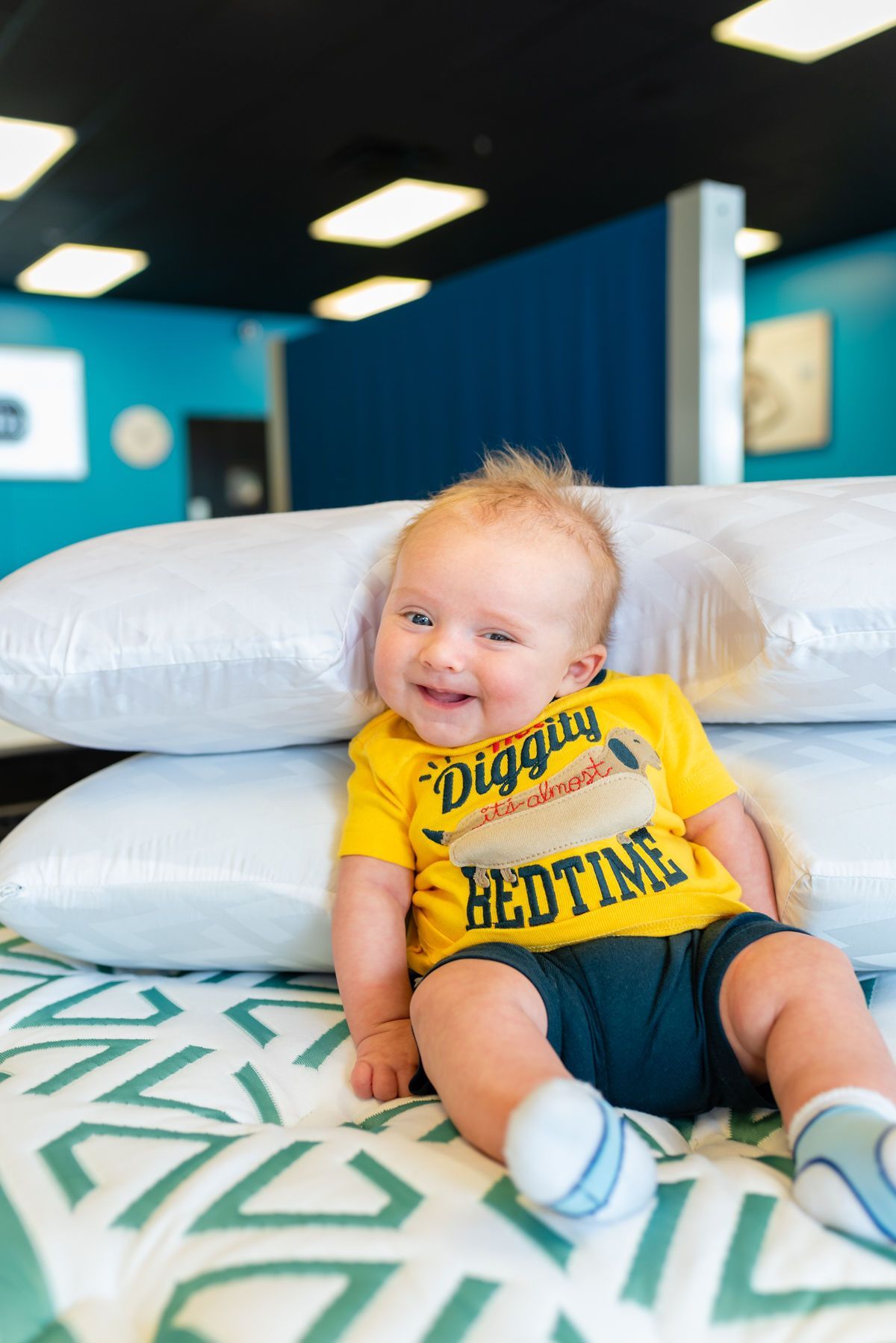 Smiling baby in a yellow shirt sitting on a bed with white pillows in a bright room