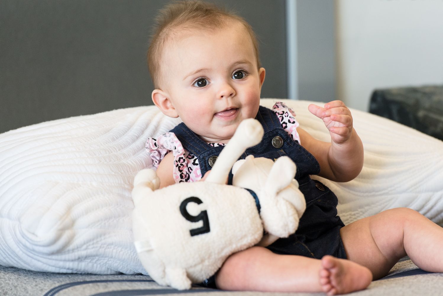 Baby sitting on a couch holding a plush toy, with a white pillow behind them.