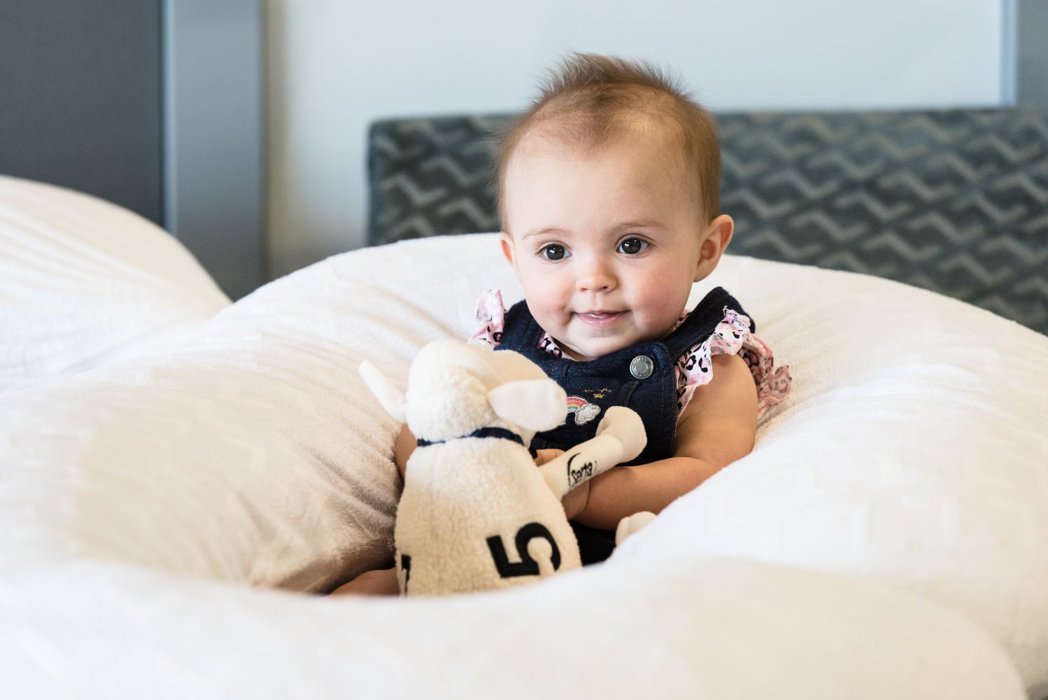 Baby sitting on a white bed, holding a stuffed toy and looking at the camera.