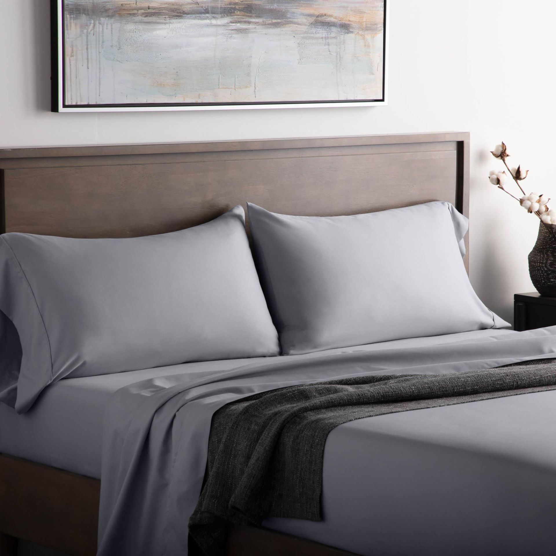 Neatly made bed with gray bedding, dark throw blanket, and a wooden headboard under abstract wall art.