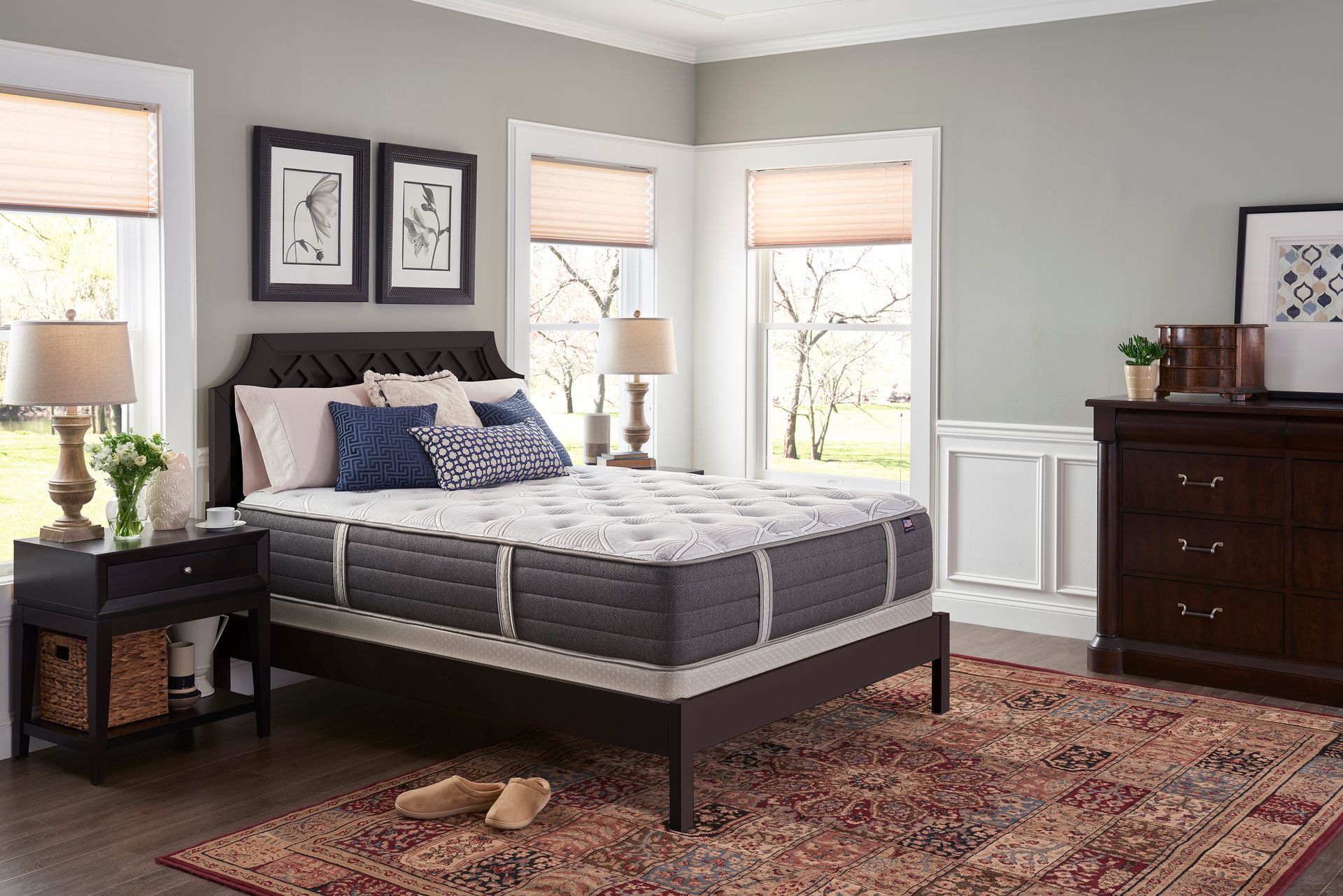 Modern bedroom with gray bed, dark wood furniture, patterned rug, and three windows with blinds.
