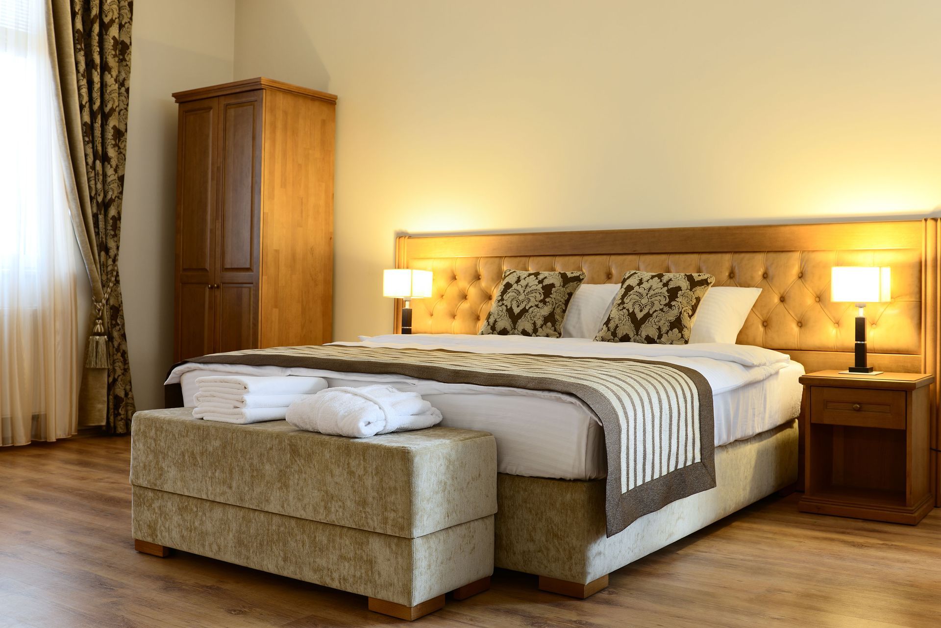 Warm bedroom with a king bed, beige bench, wooden wardrobe, and bedside lamps by a curtained window.
