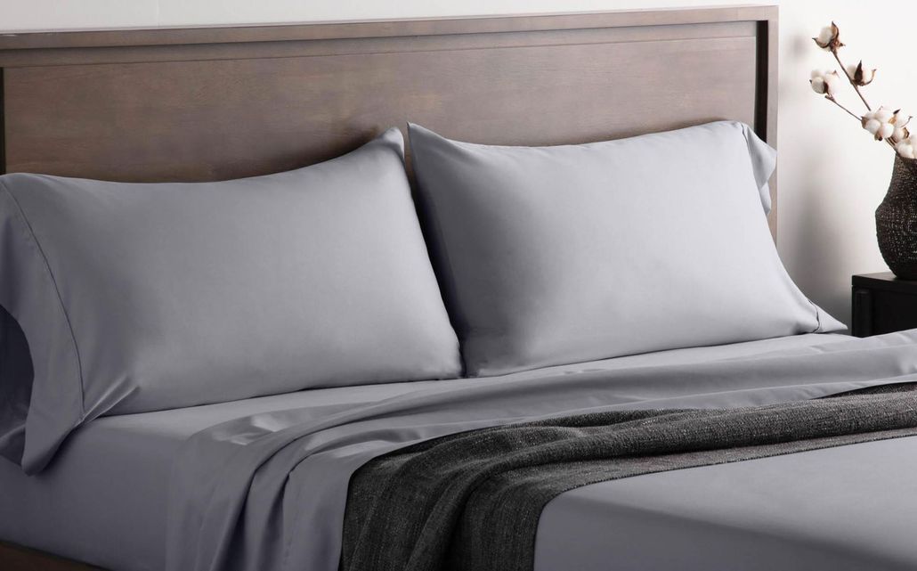 Gray bed with pillows, dark blanket, and a vase of dried flowers on a bedside table