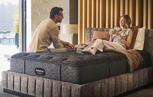 Couple sitting on a large mattress in a bright bedroom, talking and relaxing by the window.