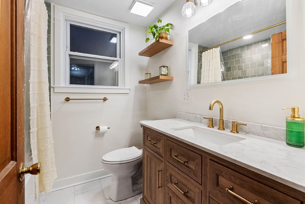 Bathroom with wood vanity, marble countertop, gold fixtures, white walls, and a window.