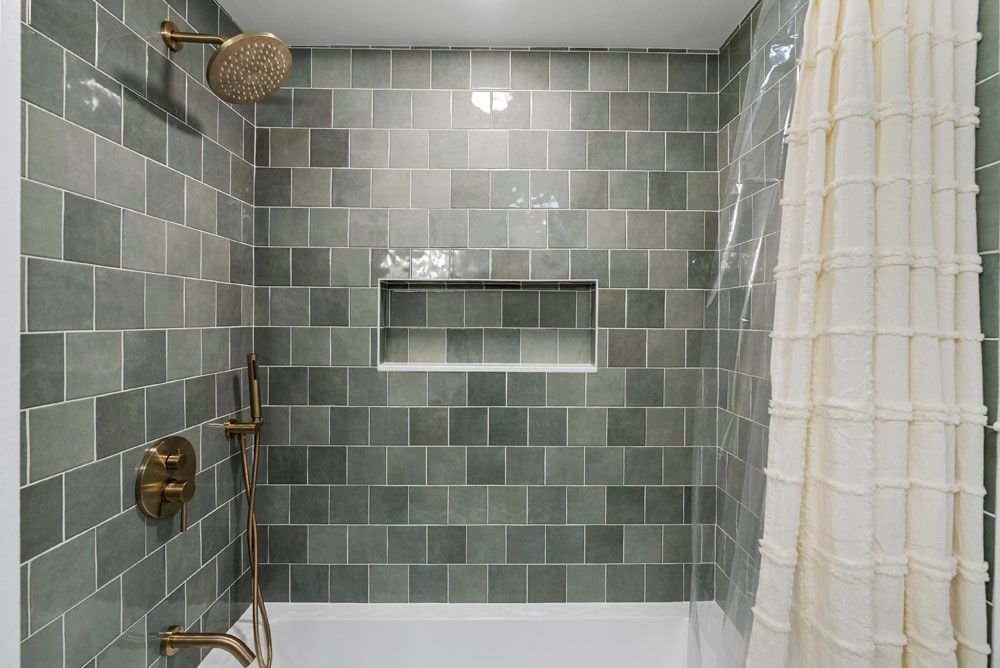 Green tiled shower with gold fixtures and a white curtain.
