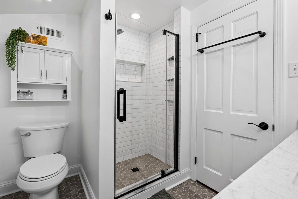 White bathroom with shower, toilet, and vanity. Black-framed shower door, white tile, and a wall-mounted cabinet.