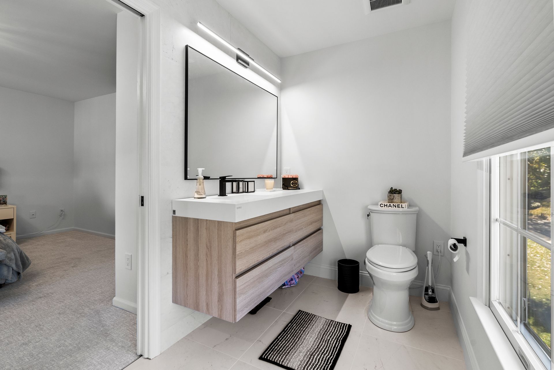 Modern bathroom with floating vanity, toilet, window, and rug. White walls and light wood accents.